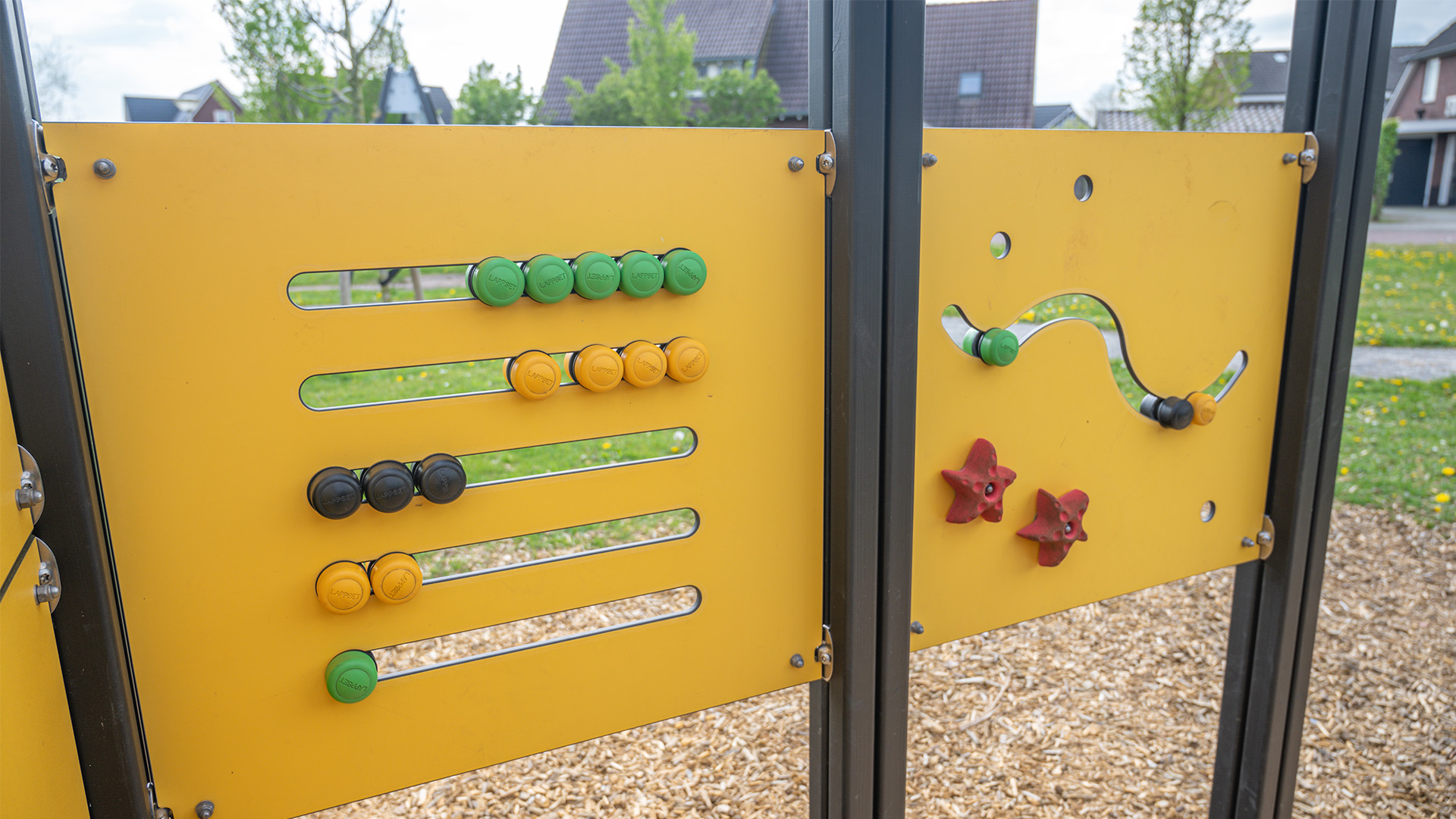 Two interactive yellow panels at a playground featuring sliding beads in various colors and a tracking game with red star-shaped handles. The ground is covered in wood chips. In the background are houses, trees, and green grass.