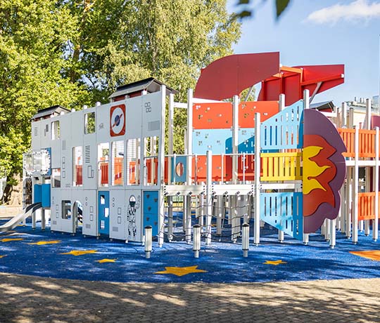 A children's playground with a space-themed climbing structure featuring rocket ship designs and colorful panels. The playground has a blue, star-patterned rubber surface and is surrounded by trees on a sunny day.