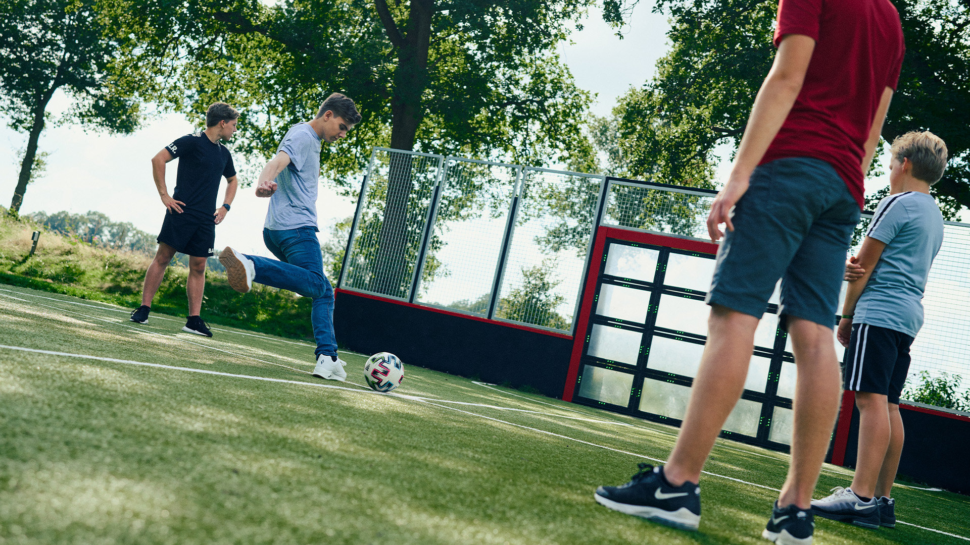 Four boys are playing soccer on a grassy outdoor field surrounded by a fence and trees. One boy in a gray T-shirt is about to kick the ball, while the other three boys watch. They are dressed in casual sportswear and appear to be enjoying the game.