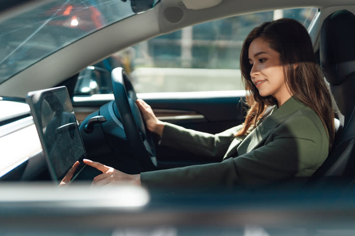 Une femme assise sur le siège conducteur d’une voiture, portant une veste verte, interagissant avec l’interface tactile du véhicule. Le visage de la personne est masqué pour préserver sa vie privée.
