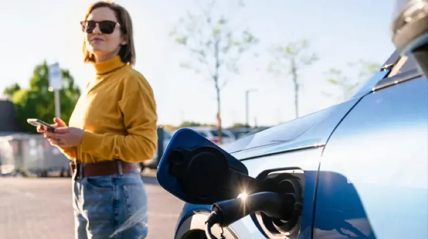 Une femme, portant des lunettes de soleil et un haut jaune, se tient à côté de sa voiture électrique bleue en cours de recharge.