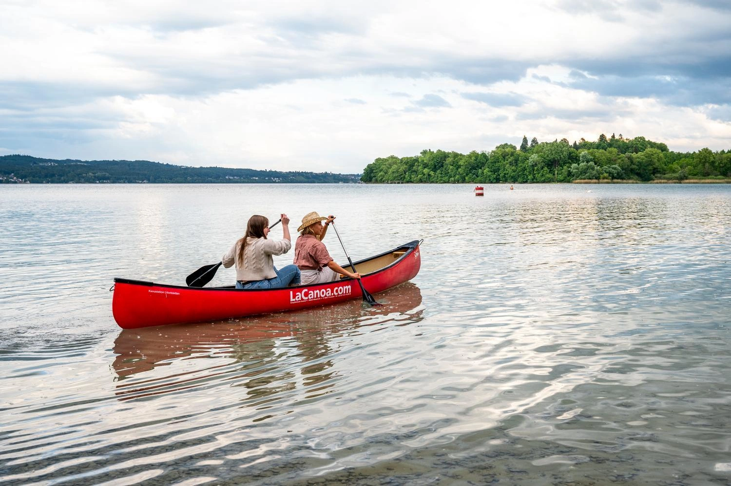 Zwei Personen paddeln in einem roten Kanu über den Bodensee, umgeben von ruhigem Wasser und grüner Uferlandschaft unter leicht bewölktem Himmel.