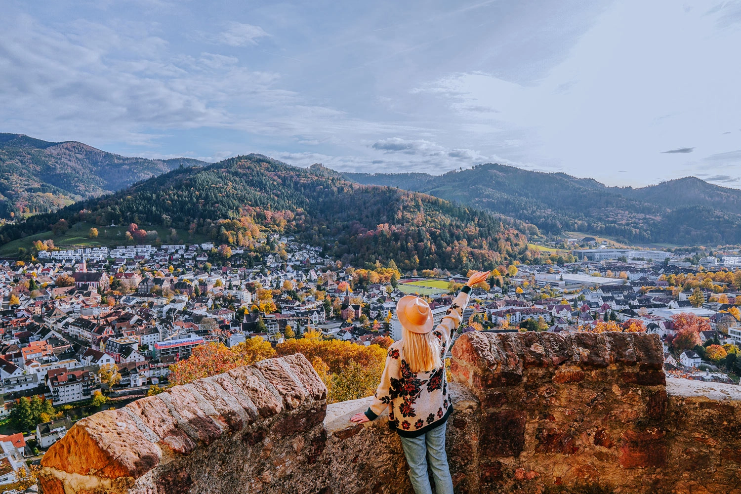 A woman looks down from a castle onto a small town surrounded by a hilly landscape of autumnal trees.