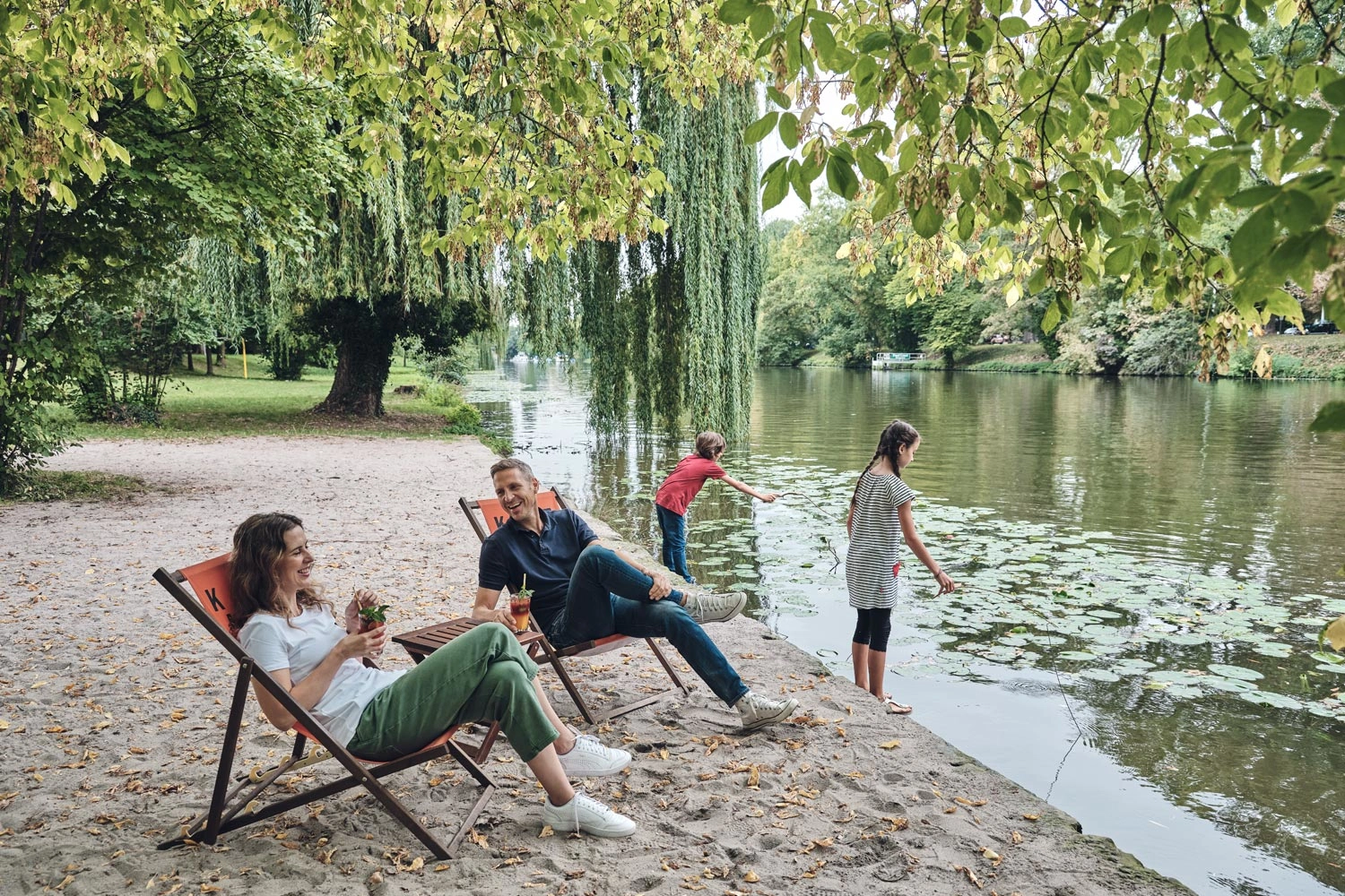 Two people are sitting in deckchairs on the sandy banks of the Neckar. Two children are playing in the river on the bank.