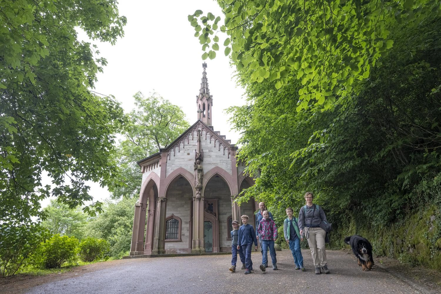 Gruppe von Wandernden mit Hund vor der historischen Klingelkapelle, umgeben von dichtem grünen Laubwald.