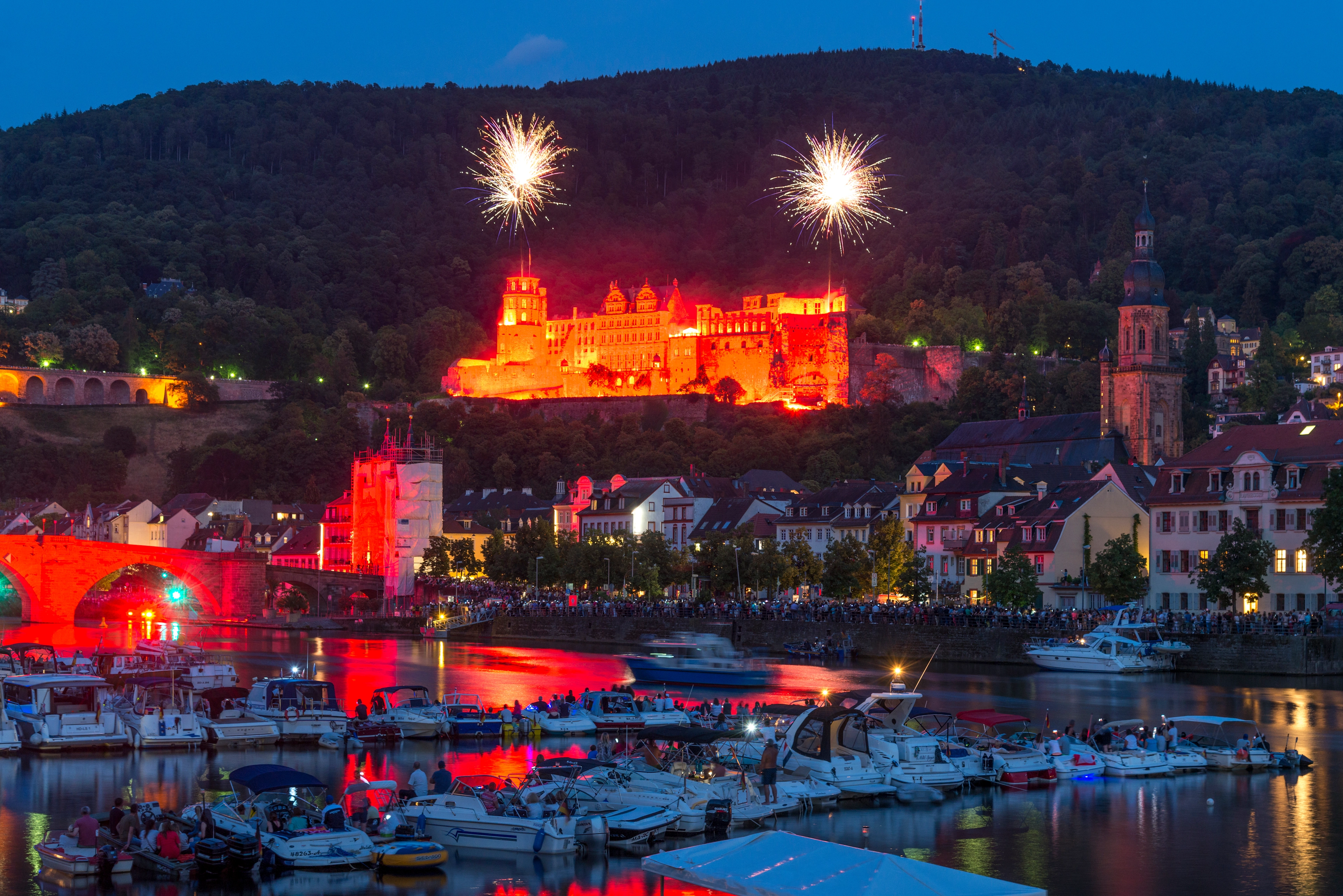 Das rot illuminierte Heidelberger Schloss während der Schlossbeleuchtung mit Feuerwerk über dem Neckar und zahlreichen Booten im Vordergrund.