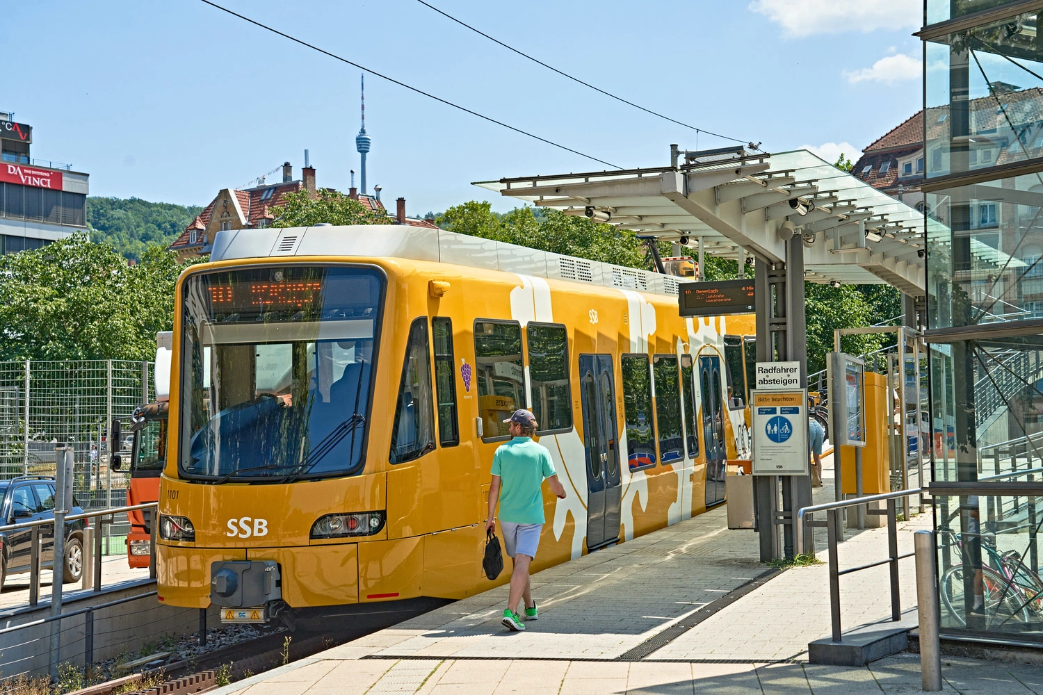 Yellow SSB light rail vehicle at the Marienplatz stop in Stuttgart, with a passenger and the television tower in the background under a blue sky.