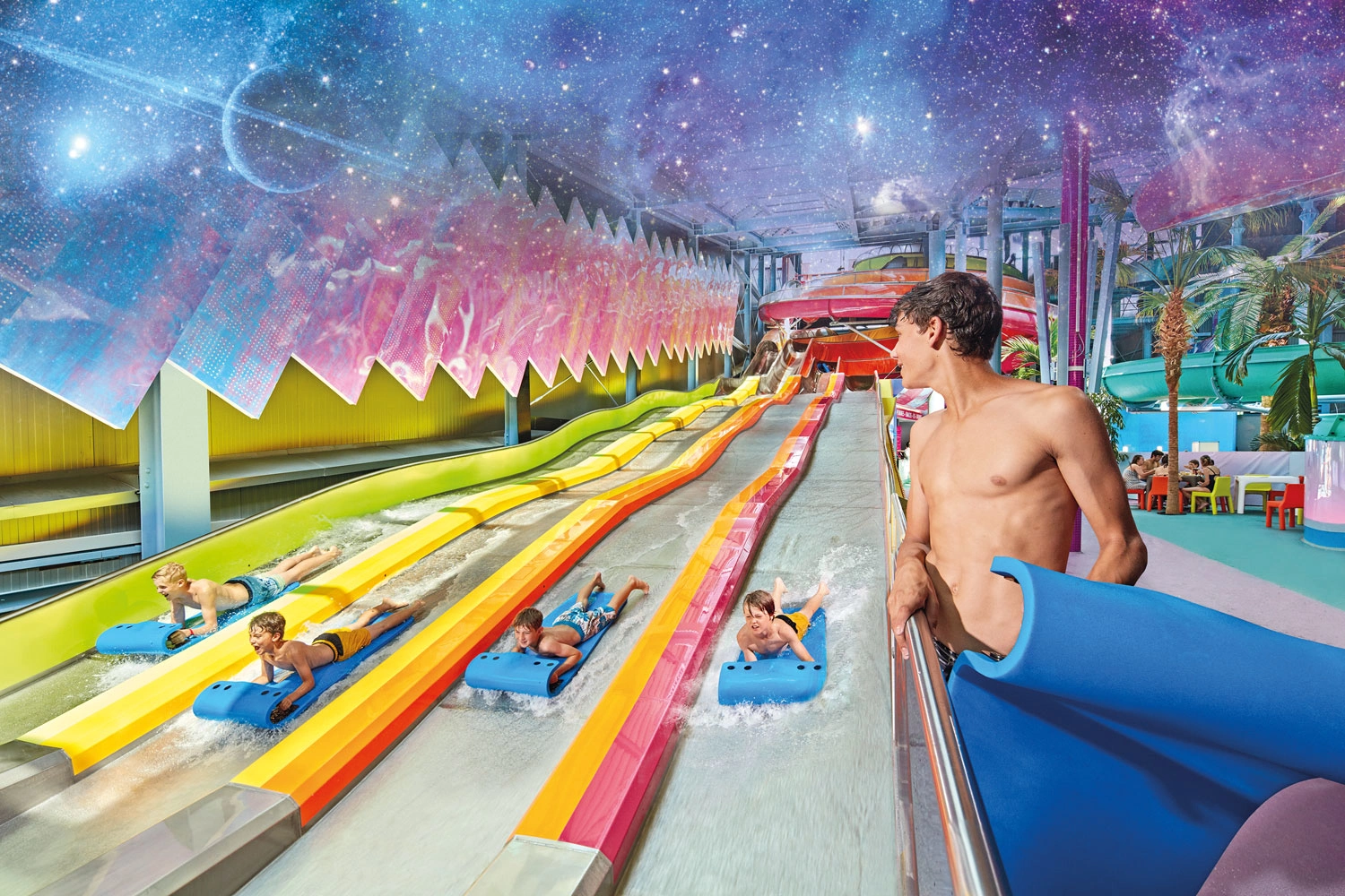 Four children are sliding down a long, colorful water slide on blue mats. A boy stands at the edge on the right and watches.