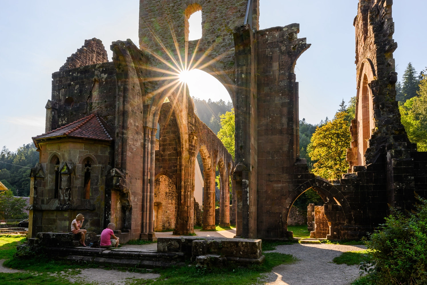 Sunlight shines through the arches of a historic monastery ruin, while two people sit in the shadow of the old walls.