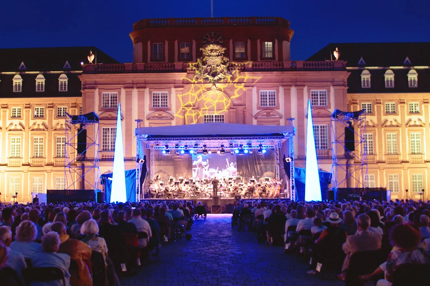 Eine beleuchtete Bühne vor einem Schloss mit sehr vielen Zuschauern. Auf der Bühne spielt ein Orchester. 