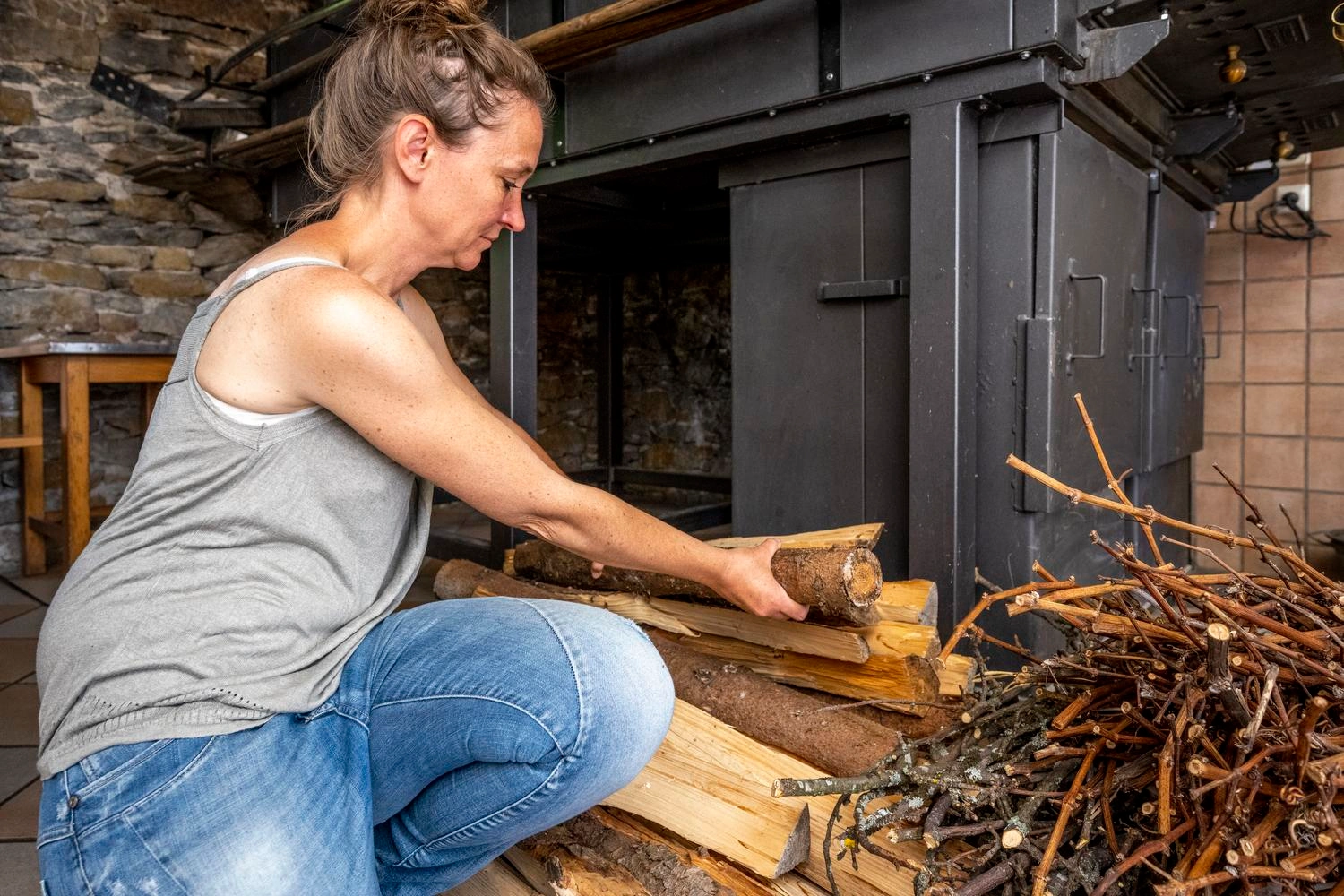 Eine Frau sitzt vor einem ofen und bereitet Holz vor, um diesen anzuheizen.