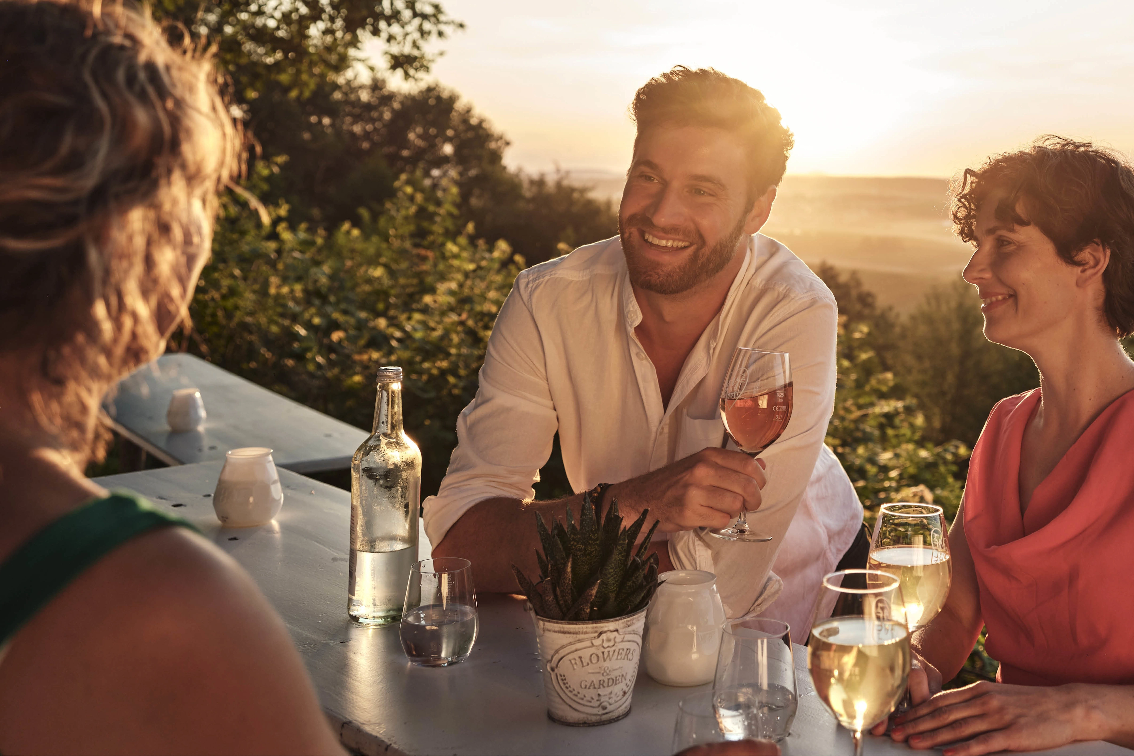Drei Personen sitzen bei Sonnenuntergang an einem Tisch mit Weingläsern und blicken über eine Landschaft; steht für geselligen Genuss und regionale Weinkultur.