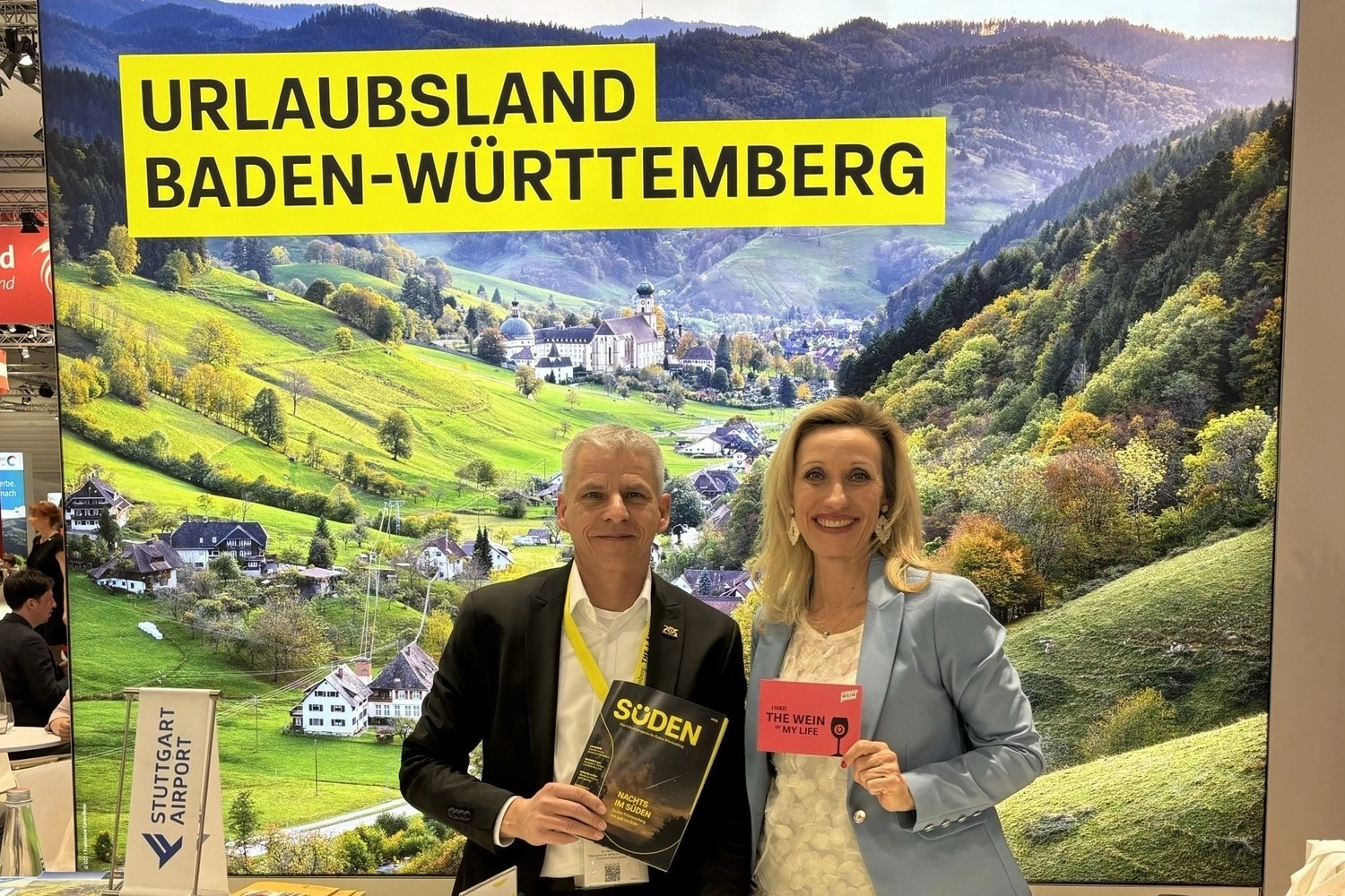 Eine Frau und ein Mann stehen vor einer großen Stellwand mit Landschaftsbild auf einer Messe.