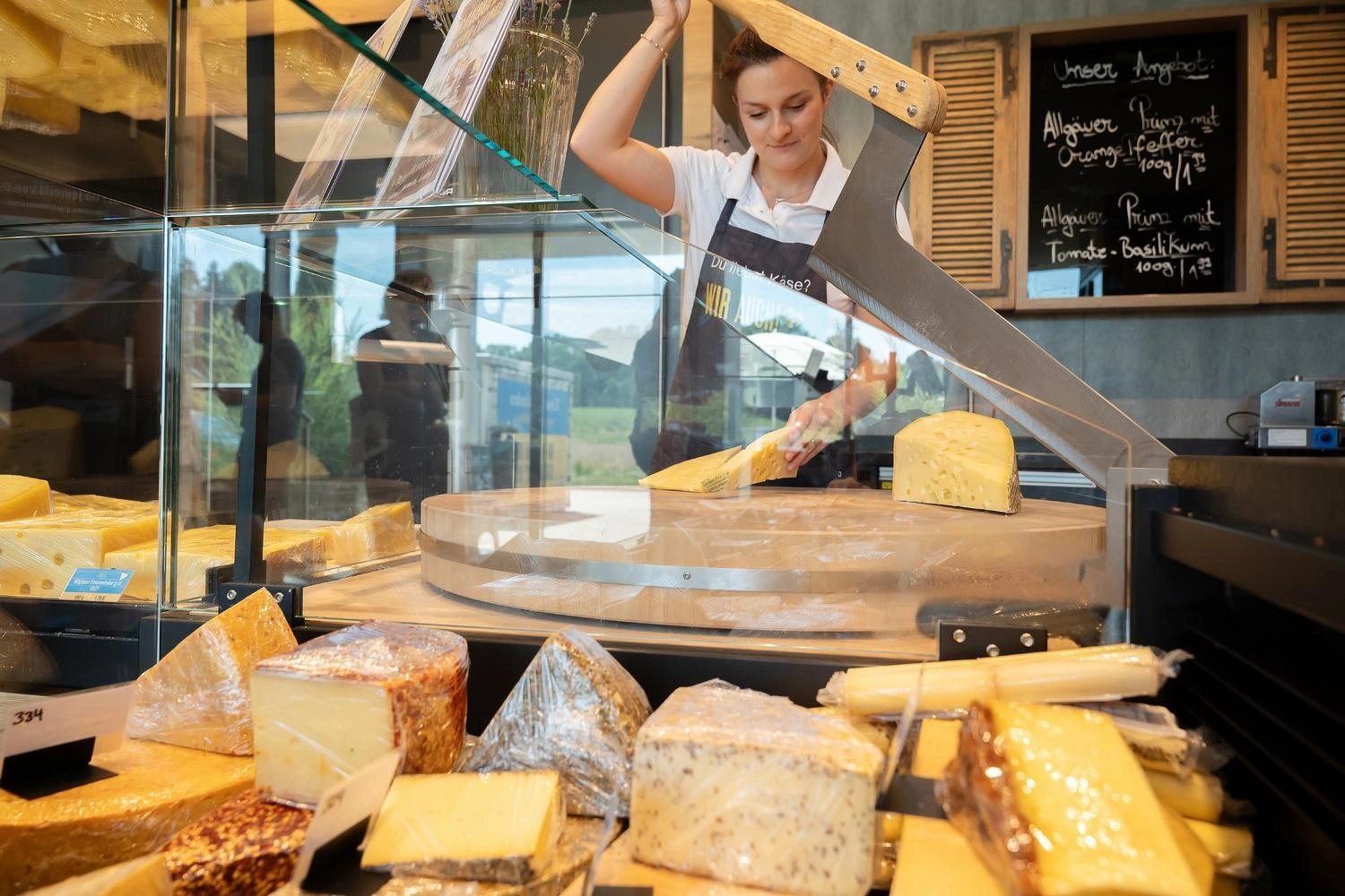 Käserei-Theke mit verschiedenen Käsesorten, während ein großes Messer ein Stück Käse auf einem runden Schneidebrett teilt.