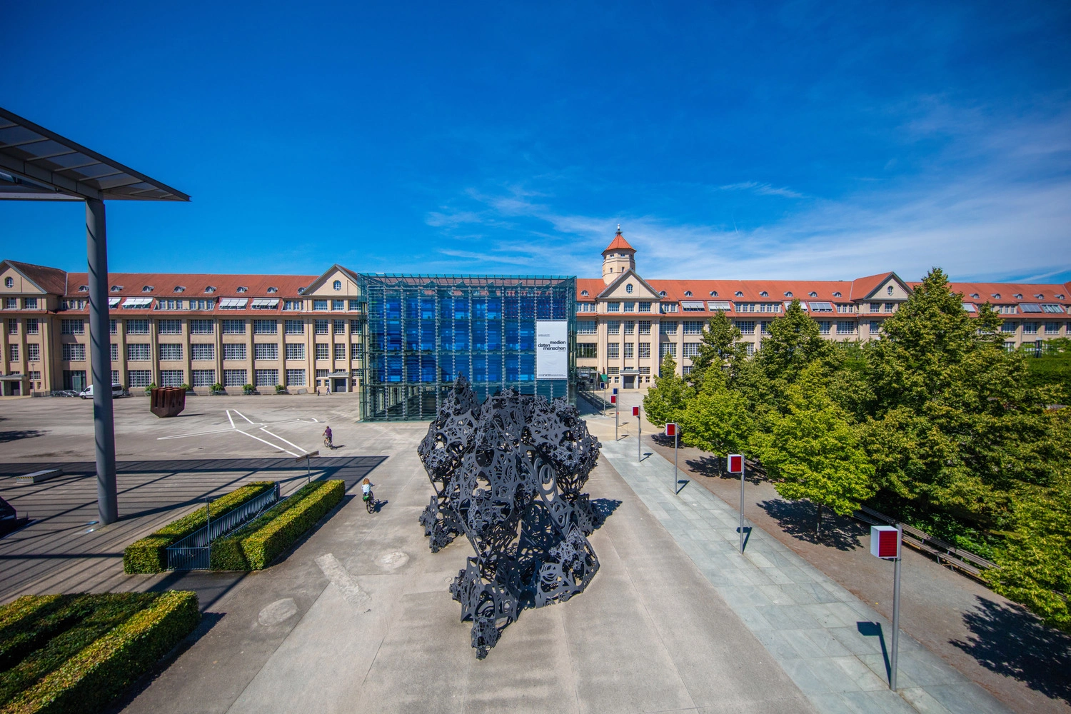 Großes Museumsgebäude mit Glasfassade und rotem Dach, davor eine moderne schwarze Skulptur auf dem Vorplatz; umgeben von Bäumen und unter blauem Himmel.