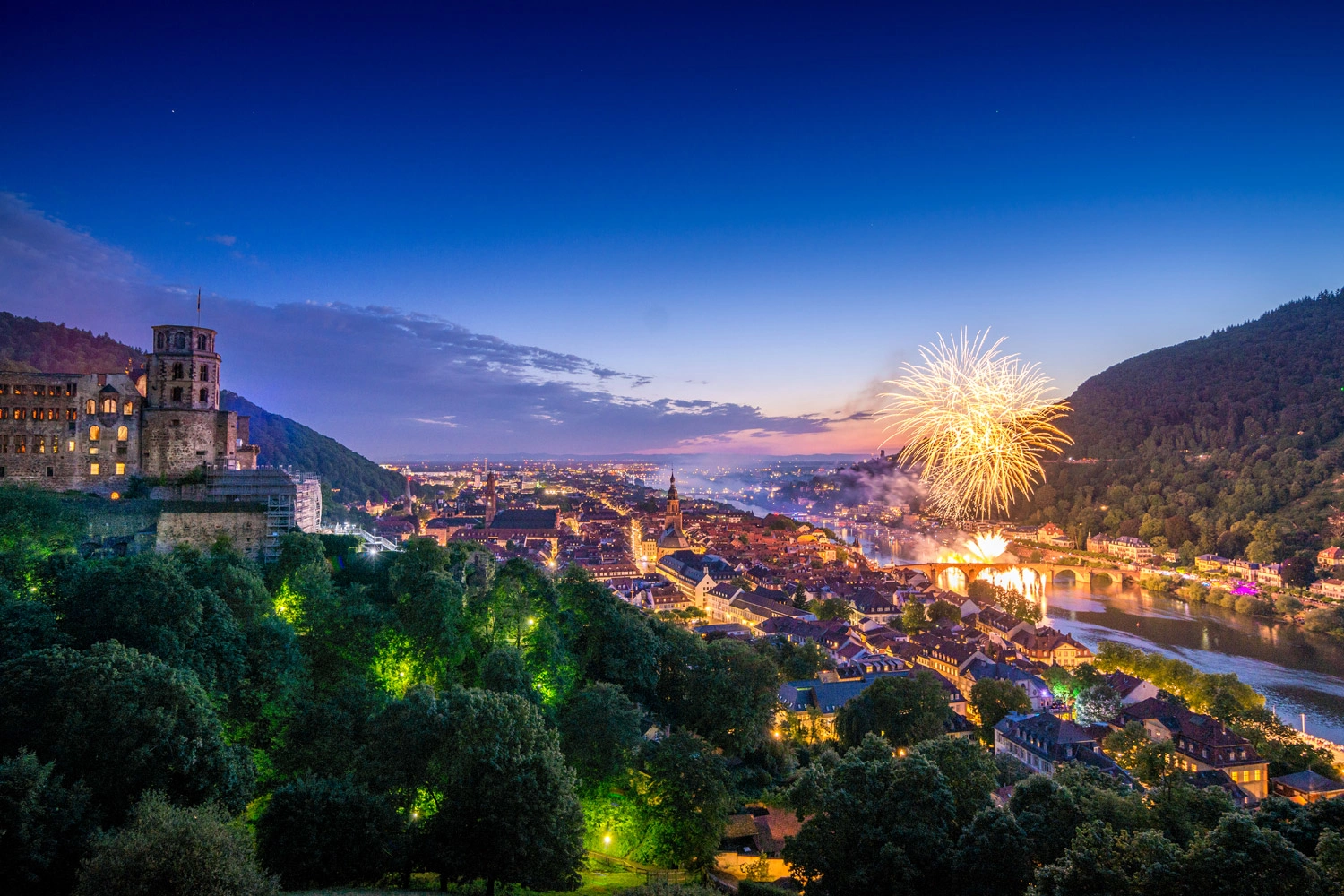 Panoramic view of a city at night, including an illuminated castle. A large fireworks display can be seen over the river on the right-hand side.