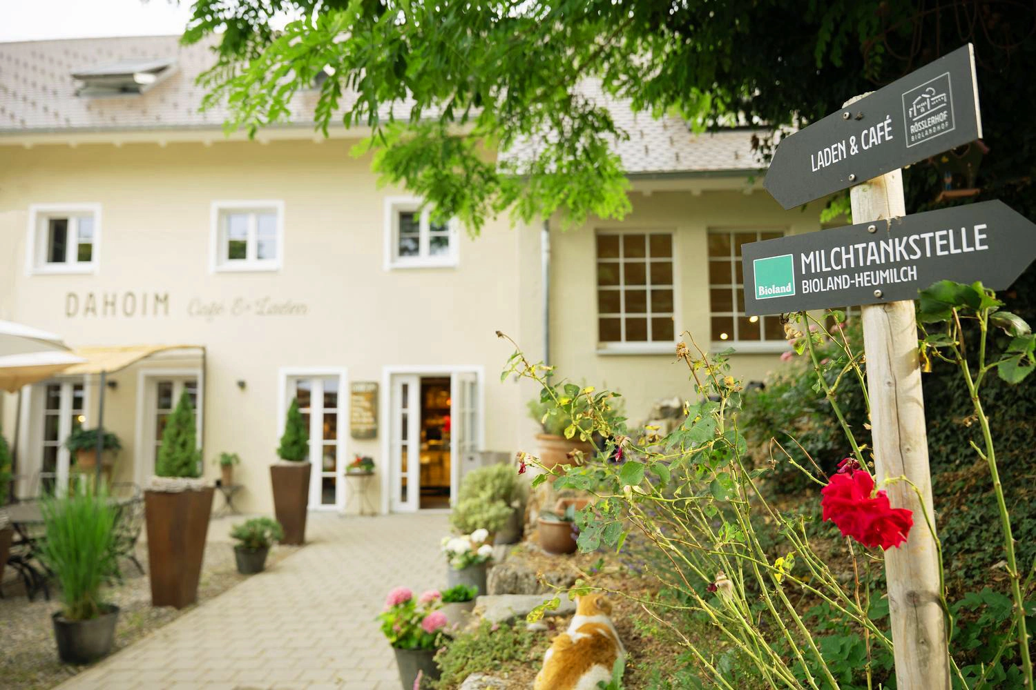 Eingang eines Hofcafés mit Blumenschmuck und Wegweisern zu Laden und Milchtankstelle unter grünem Baum.