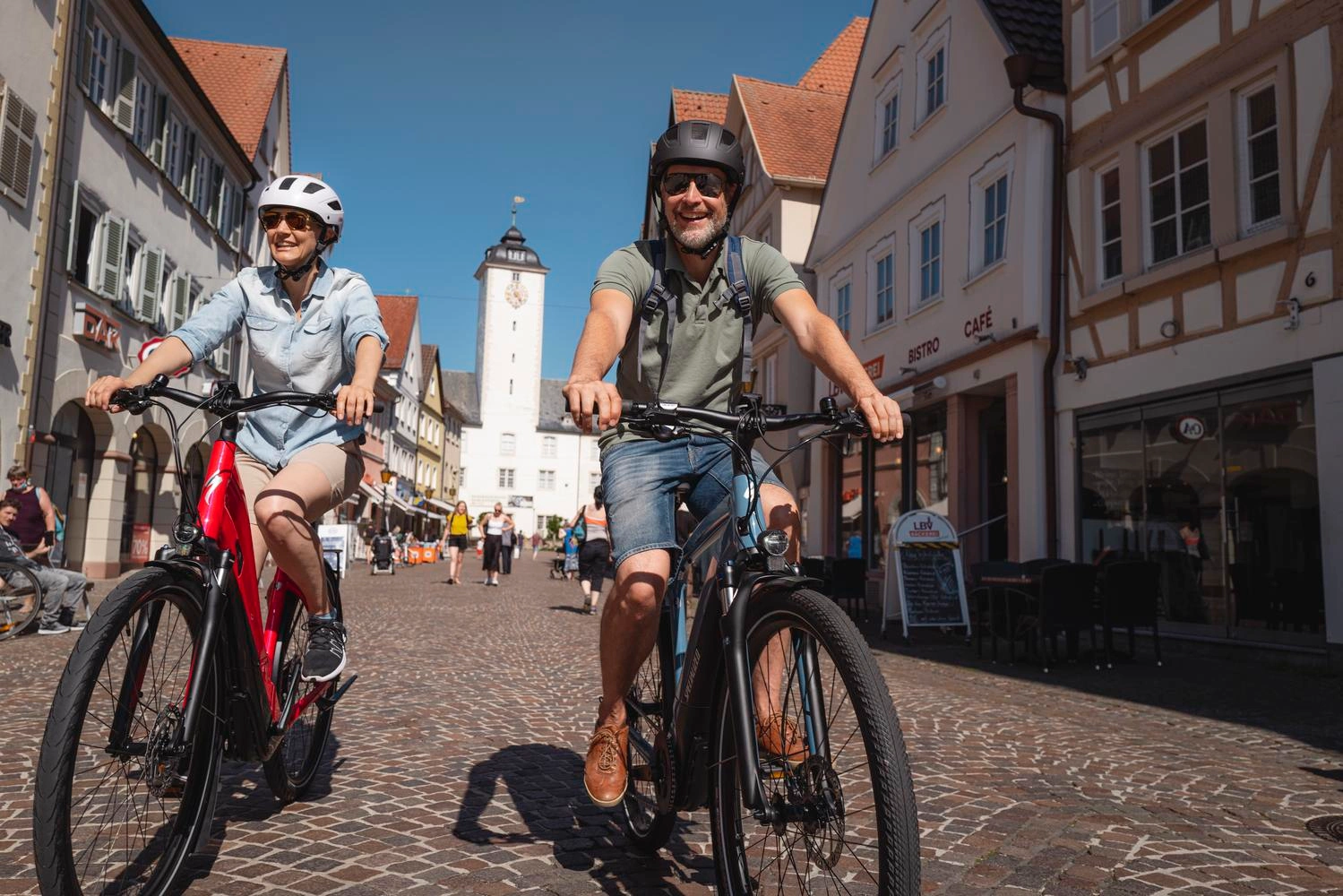 Zwei Radfahrer fahren durch eine Altstadt. Im Hintergrund ist ein Kirchturm zu sehen.