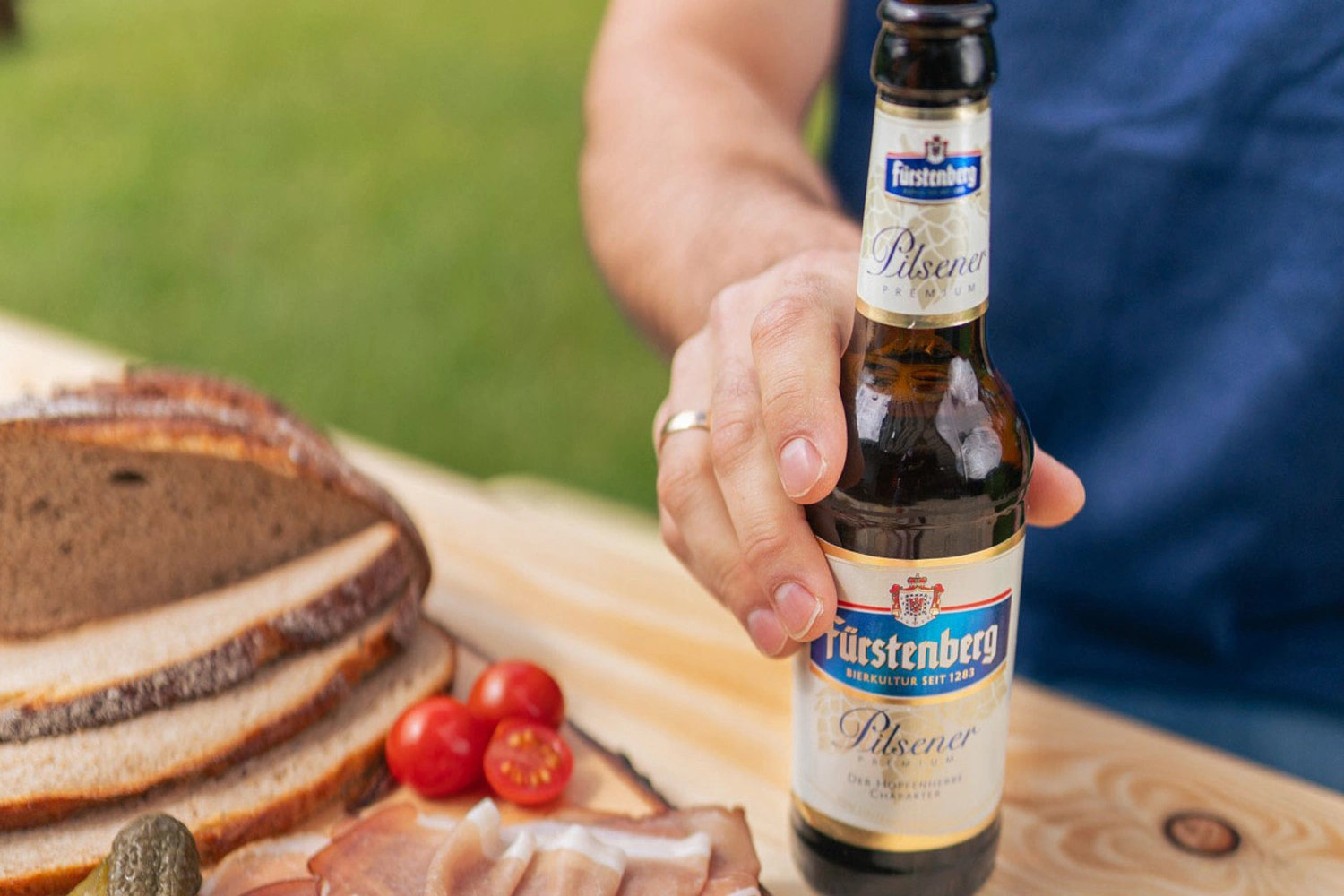 One person is holding a bottle of beer in one hand. Next to him is a snack board with bread and ham.
