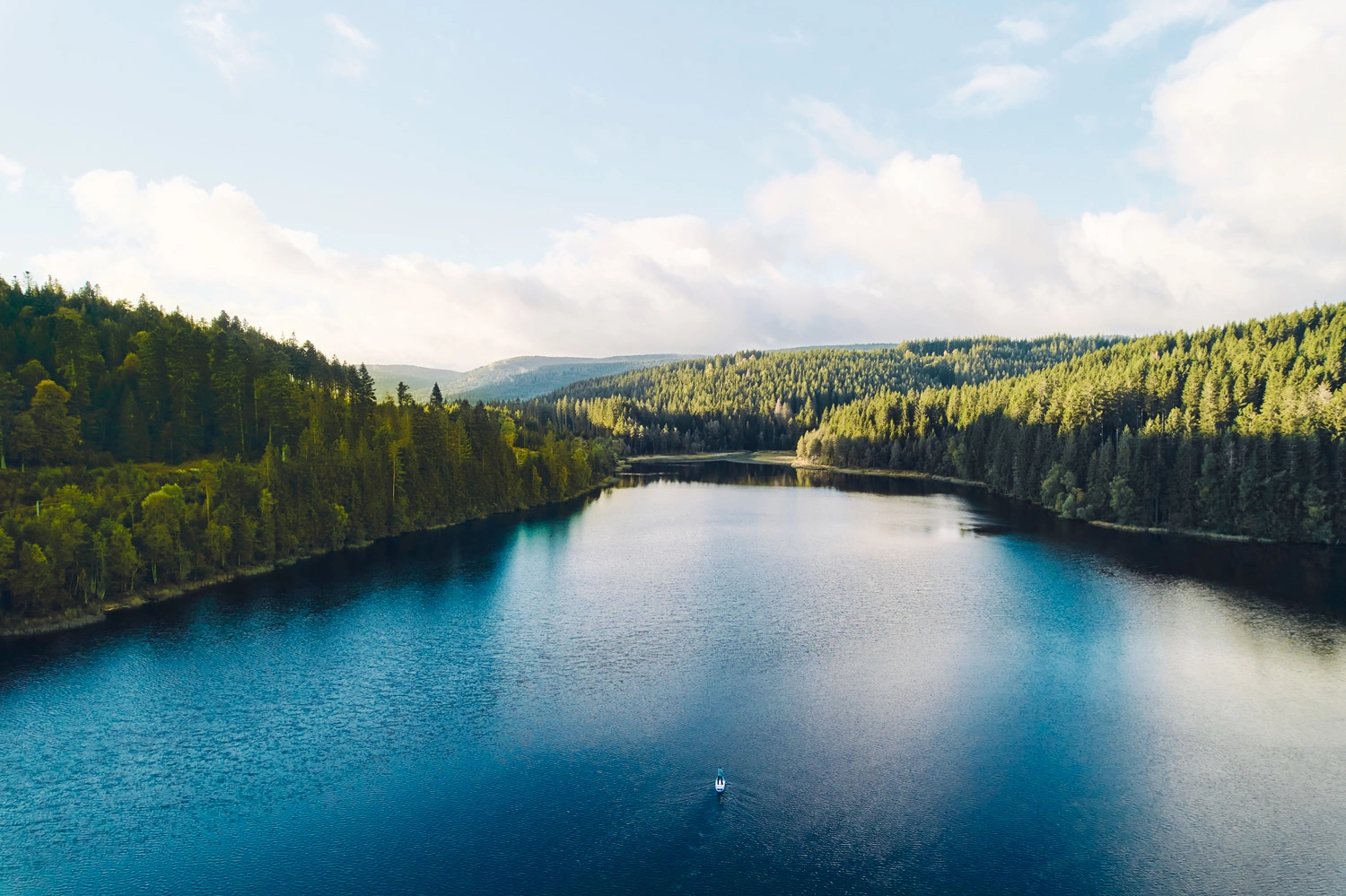 Large, tranquil lake nestled between densely wooded hills; the water reflects the sky as clouds drift across the vast landscape.