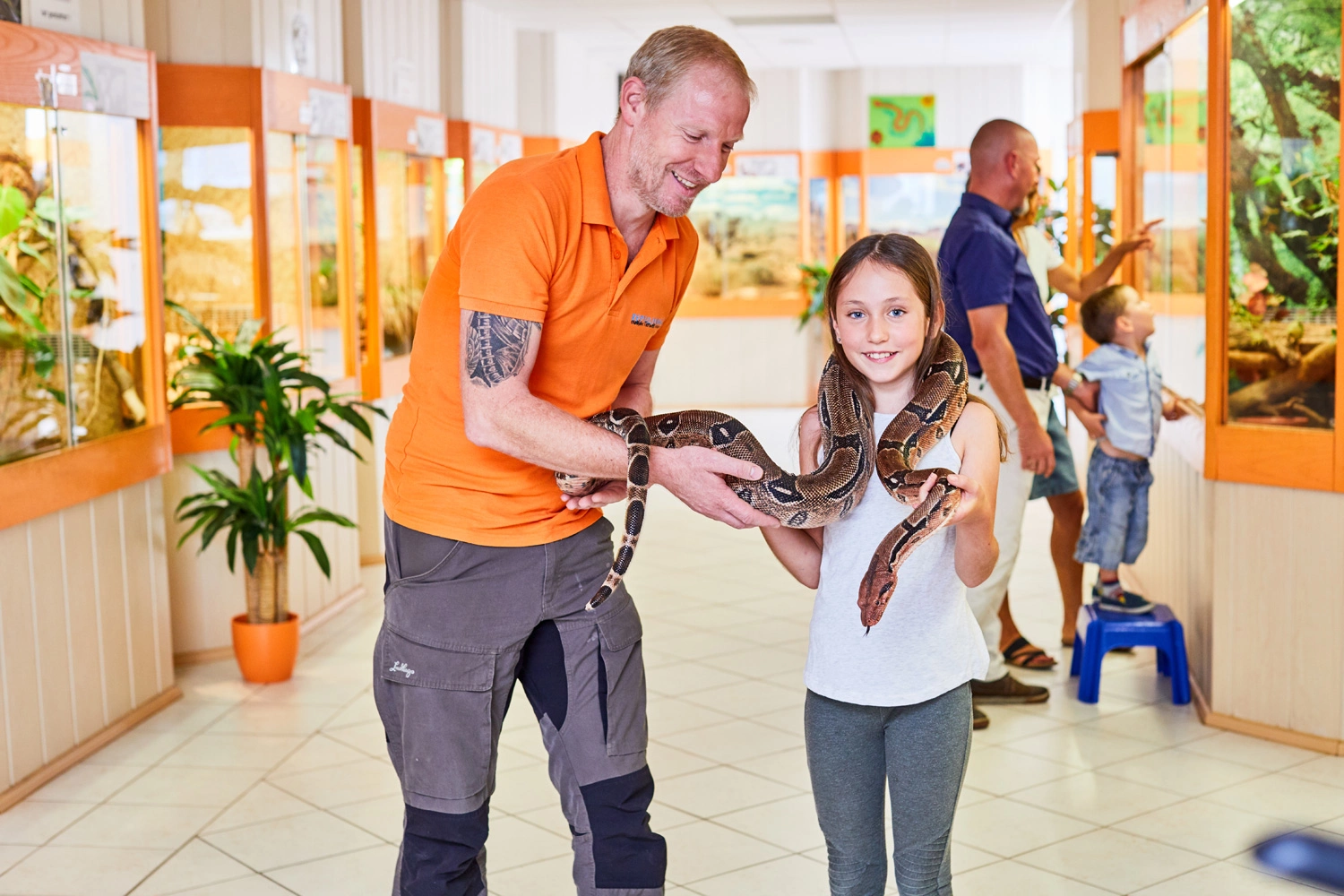 Ein Mädchen hält in einem Reptilienhaus eine Schlange auf ihren Schultern. Ein Tierpfleger hält die Schlange dabei fest.