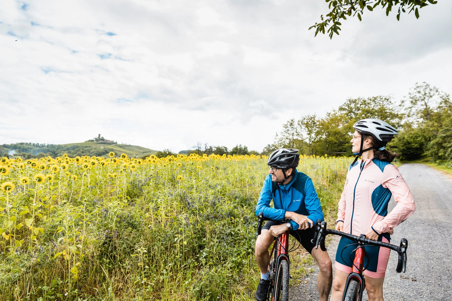 Ein Radfahrer lehnt auf seinem Lenkter. Neben ihm steht eine Radfahrerin. Sie blicken beide über ein Sonnenblumenfeld. Am Horizont steht eine Burg auf einem Berg. 
