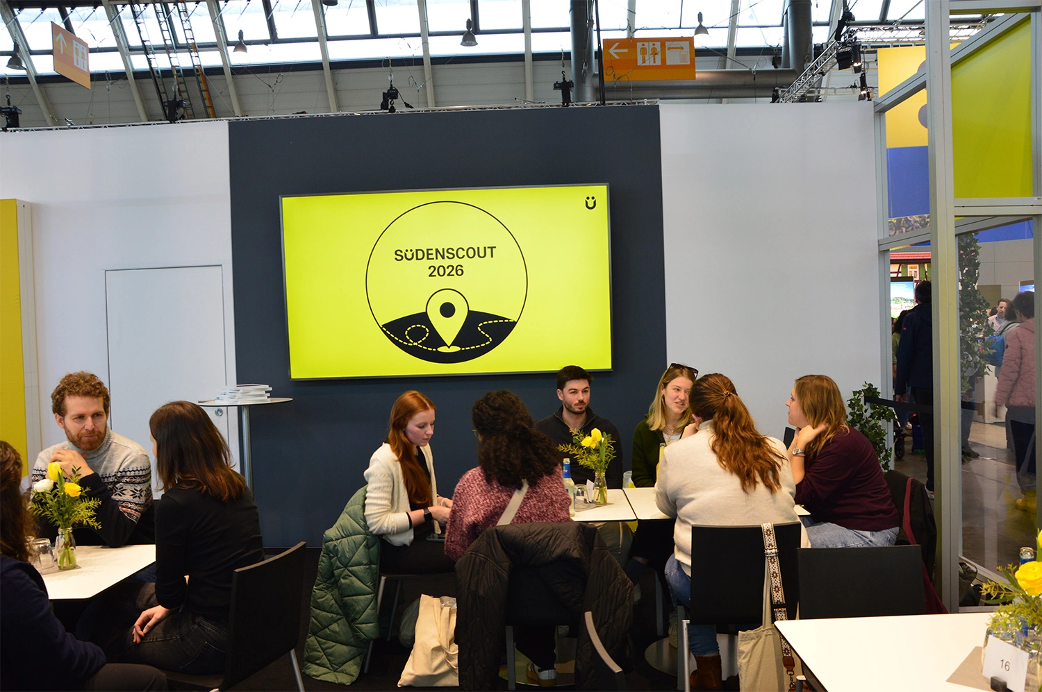 Ein Messestand mit einer großen gelben „Sudenscout 2026“-Anzeige, an dem mehrere Personen an Tischen sitzen und sich austauschen.