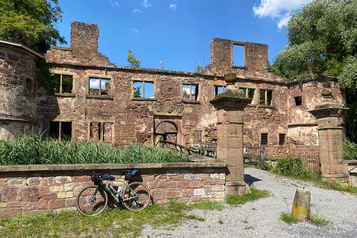 Ruins of a historic sandstone building with open windows and collapsed walls; in front of it, a bicycle parked at the entrance gate.