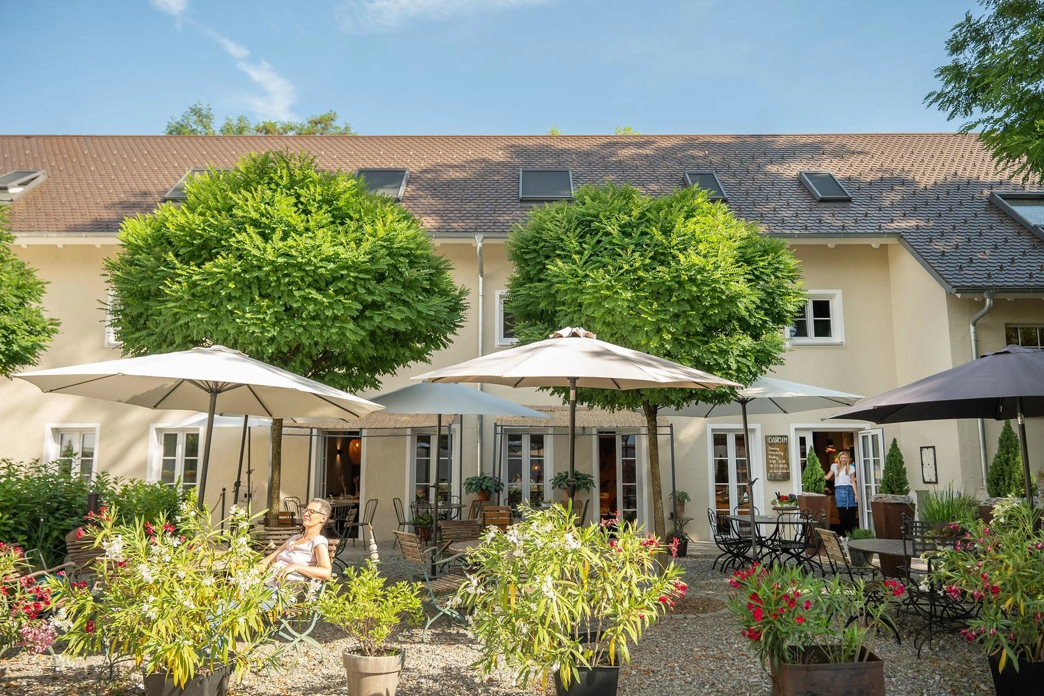 Gemütlicher Innenhof eines Hofcafés mit Sonnenschirmen, Holztischen und üppigen Pflanzen vor einem hellen Gebäude.