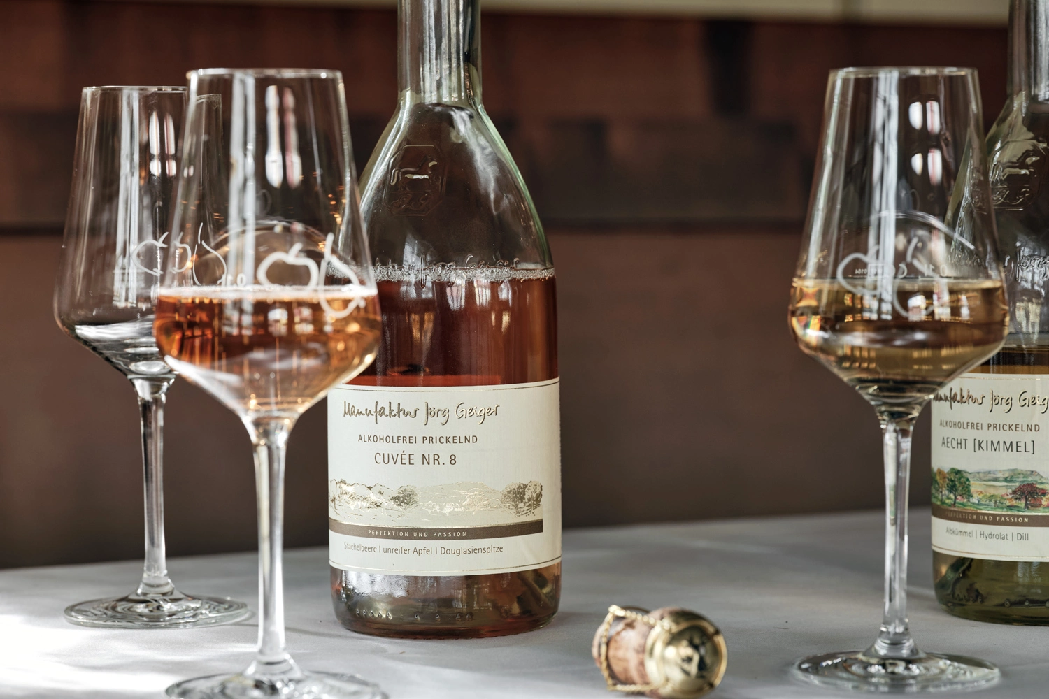 Flaschen und Gläser mit prickelndem alkoholfreiem Cuvée von der Manufaktur Jörg Geiger auf einem Tisch, daneben ein Flaschenkorken – edle Getränke in stilvollem Ambiente.