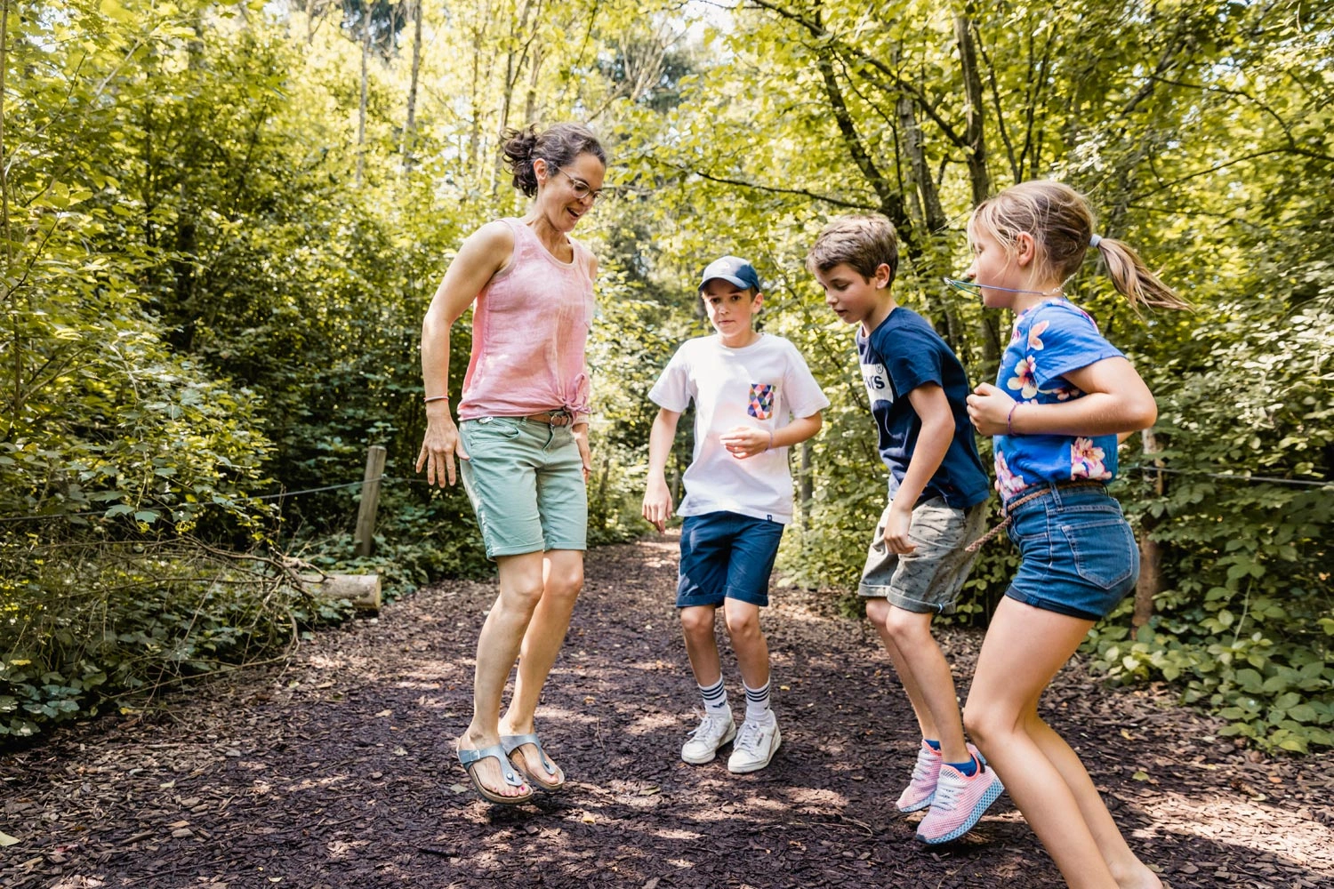 Eine Frau springt mit ihren drei Kindern auf einem Naturtrampolin auf einem Waldboden herum.