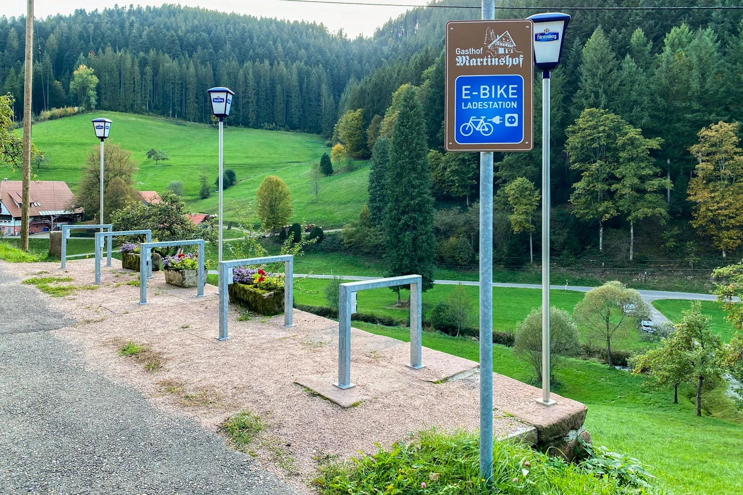 E-An e-bike charging station in the Black Forest, surrounded by green meadows and wooded hills, with several bicycle stands nearby.
