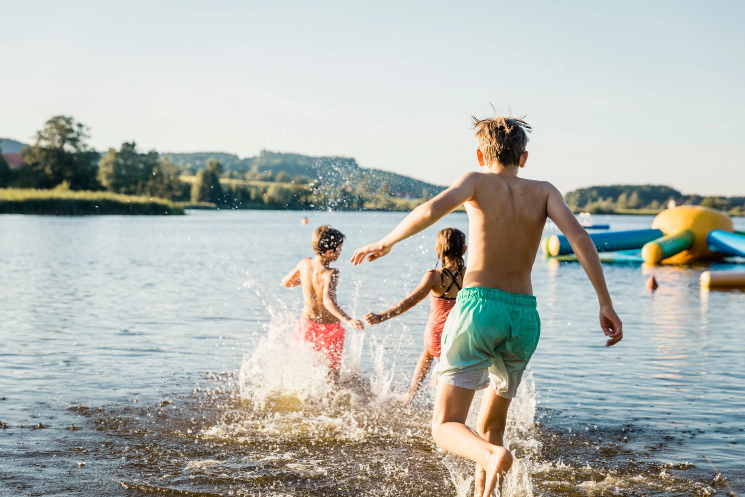 Drei Kinder rennen bei strahlendem Sonnenschein vergnügt ins Wasser eines Sees.