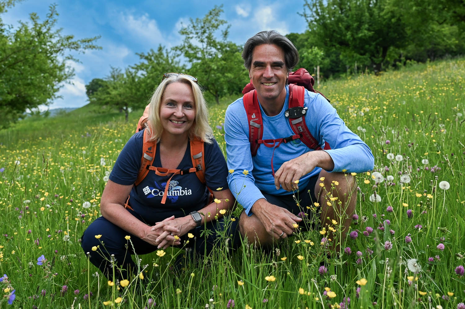 Eine Frau und ein Mann in Wanderklamotten knieen in einer Blumenwiese und schauen in die Kamera.