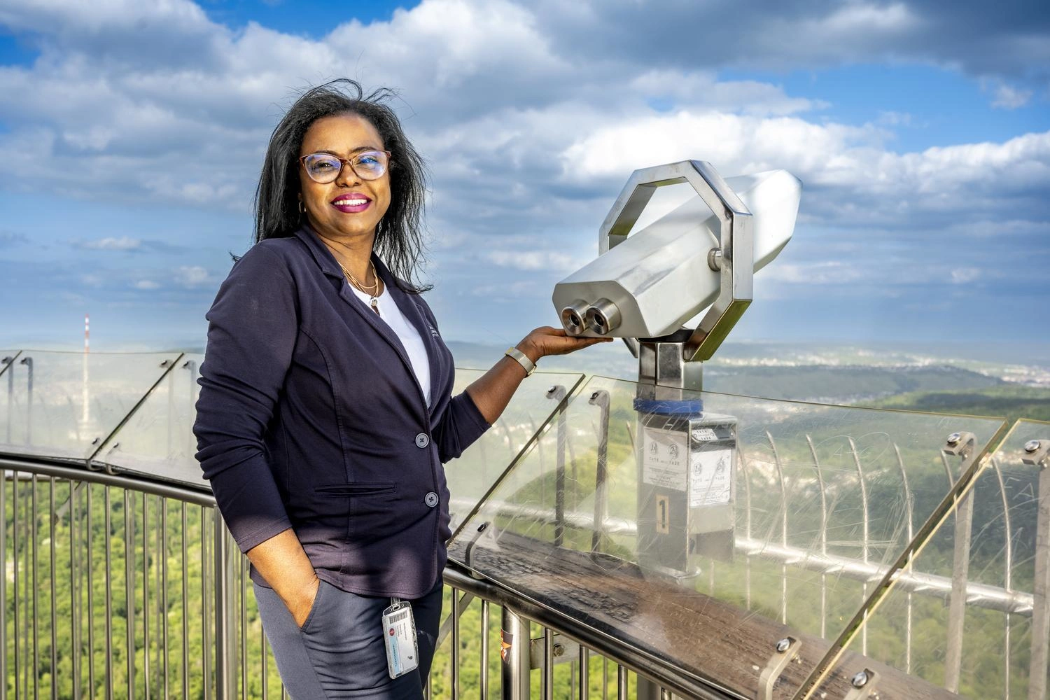 Person steht auf der Aussichtsplattform des Stuttgarter Fernsehturms neben einem silbernen Fernglas, mit Blick auf Wälder und Stadt unter blauem Himmel mit Wolken.