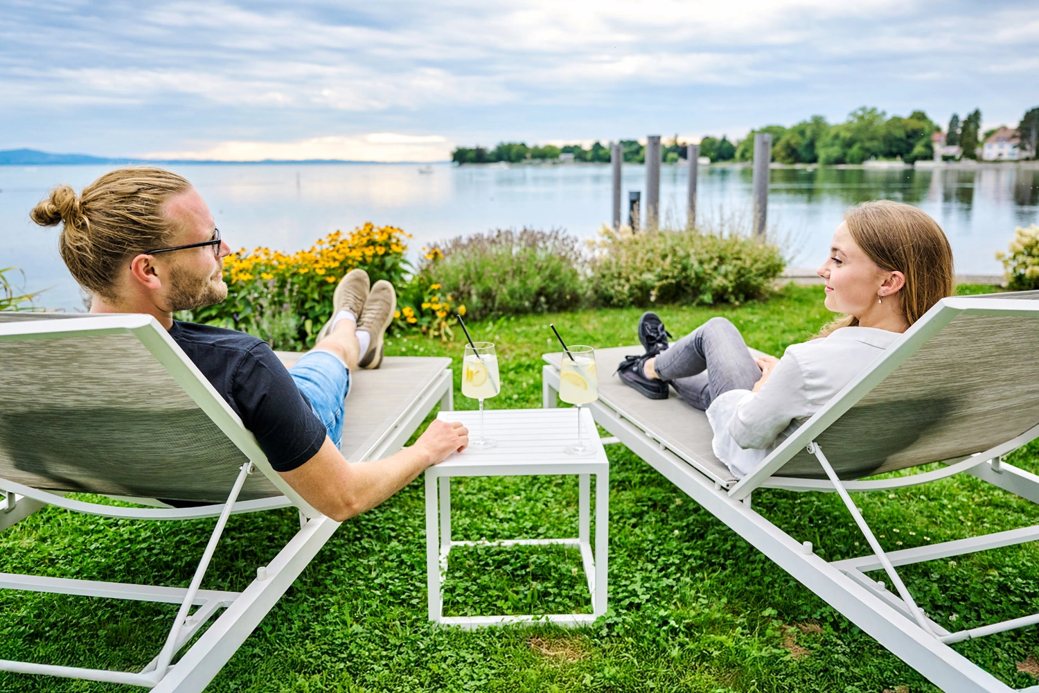 Two people relax on deck chairs by the lake, in front of them a small table with drinks, in the background calm water, flowers, and a cloudy sky.