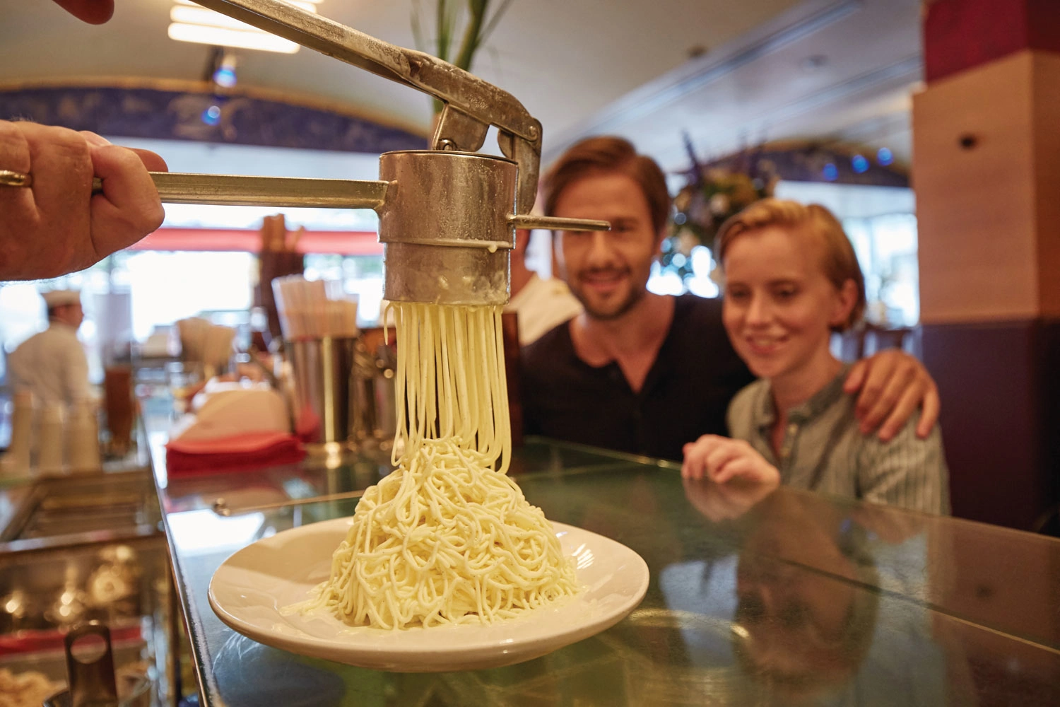 Vanilleeis wird auf einer Theke durch eine Presse zu dünnen Schnüren gepresst. Hinter der Theke warten freudig zwei Personen auf das frisch zubereitete Spaghettieis.