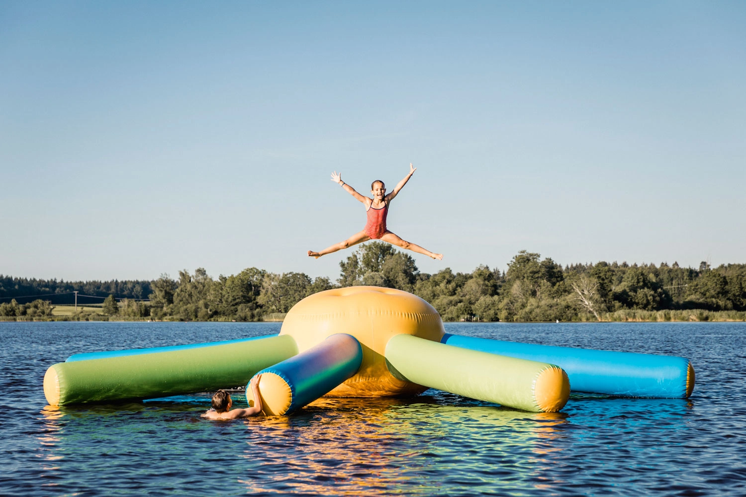 In der Mitte eines Sees liegt eine große Schwimmmatratze in Form einer Krake. Ein Mädchen springt darauf in die Höhe.