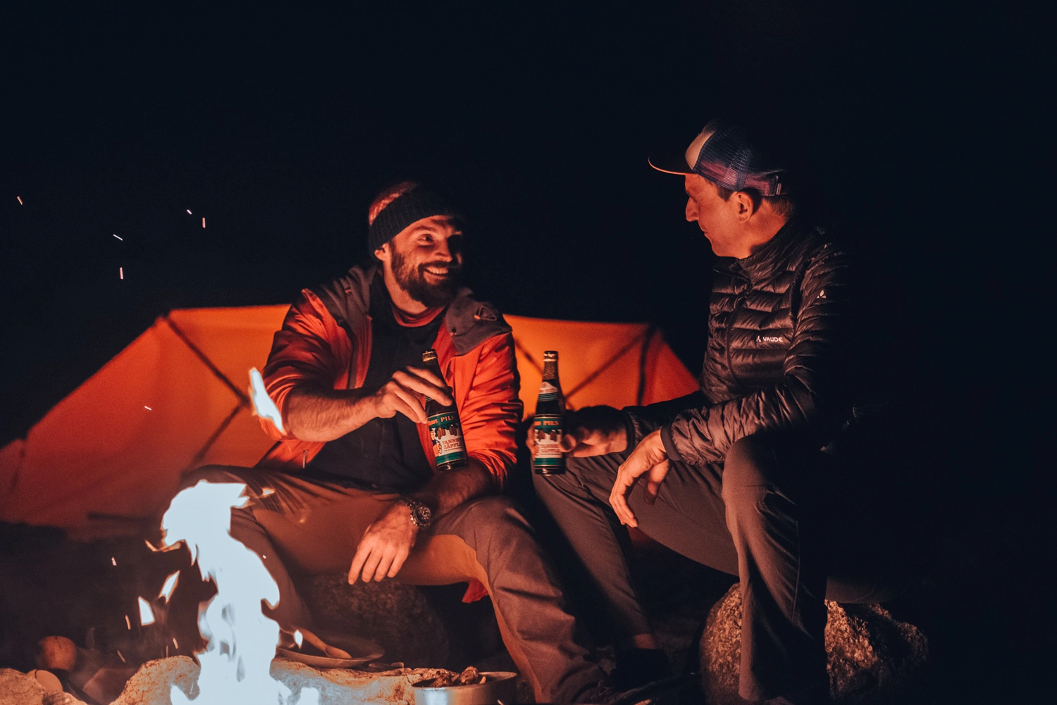 Two men are sitting around a campfire at night, toasting each other with a bottle of beer.