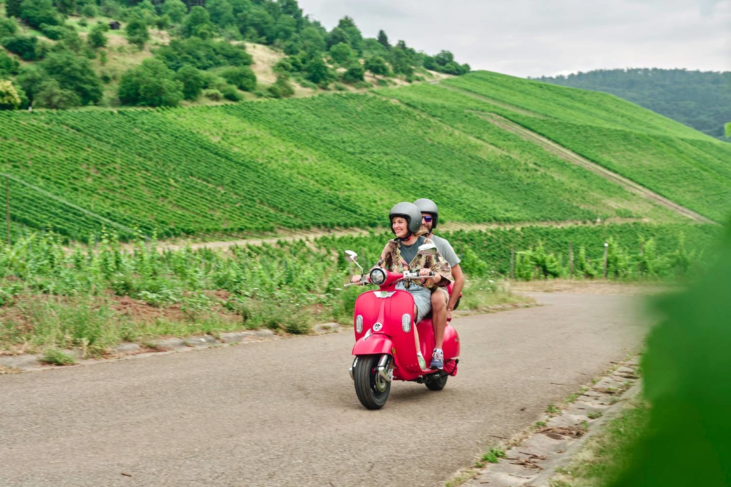 Personen fahren auf einer roten Vespa durch Weinberge im Remstal. Geschwungene Rebhänge und grüne Landschaft prägen die Weinregion Baden-Württembergs.