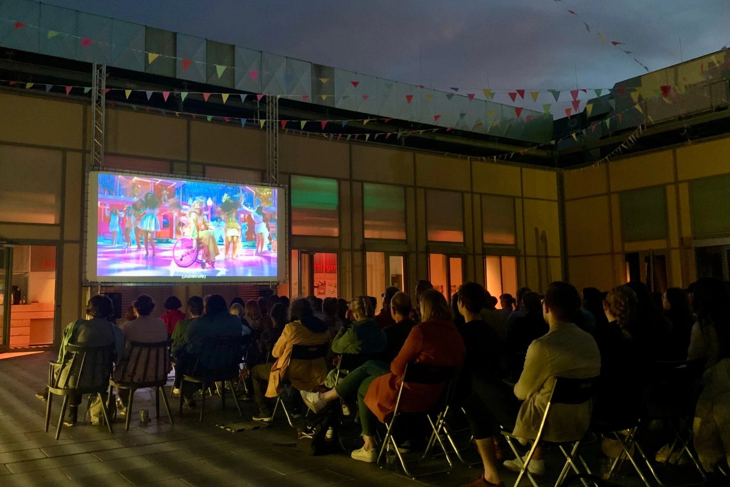 A cinema screen is set up in the courtyard of a building. People sit in front of it and watch a film under the open night sky.