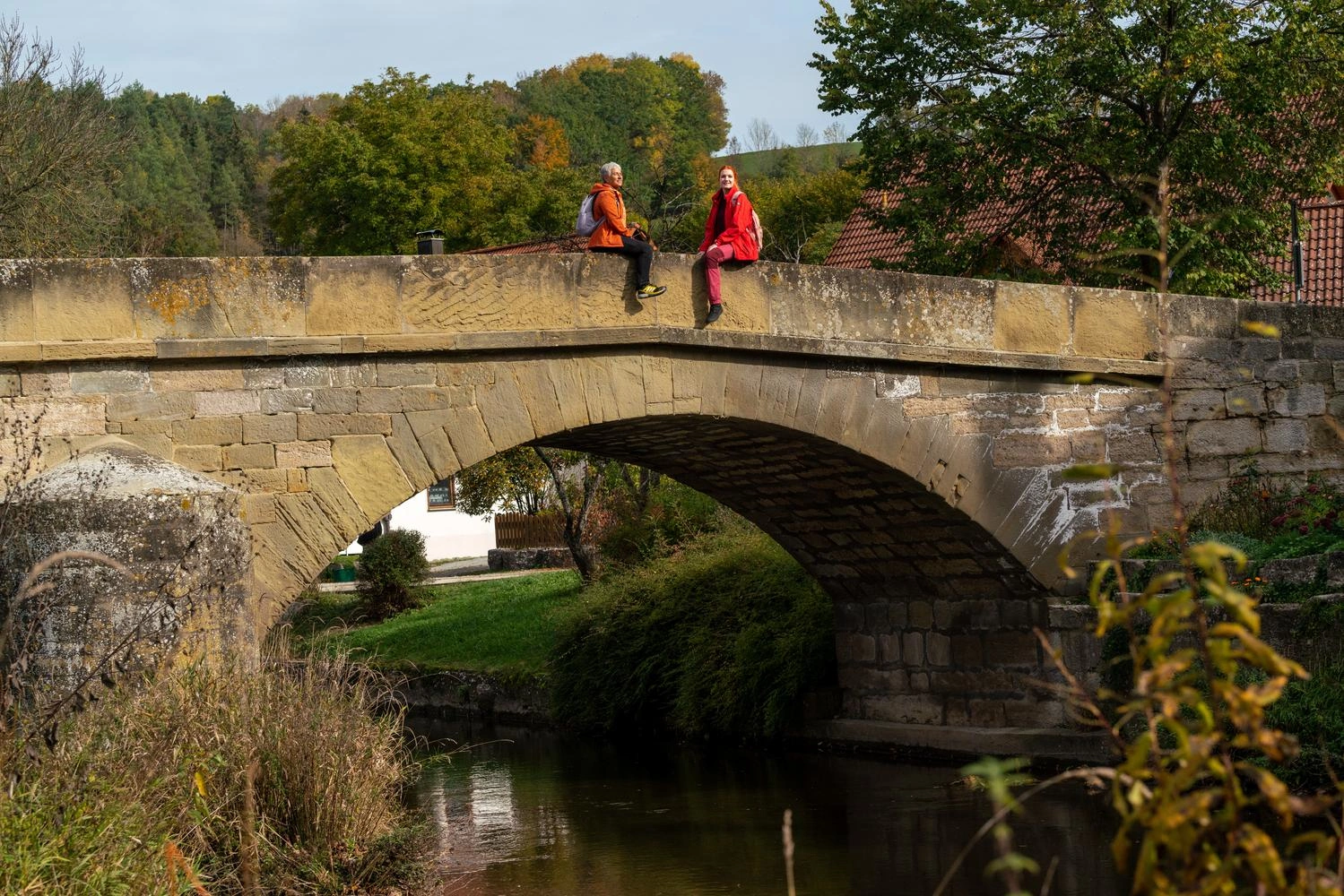  Two hikers take a break and sit on a stone bridge in the evening sun.