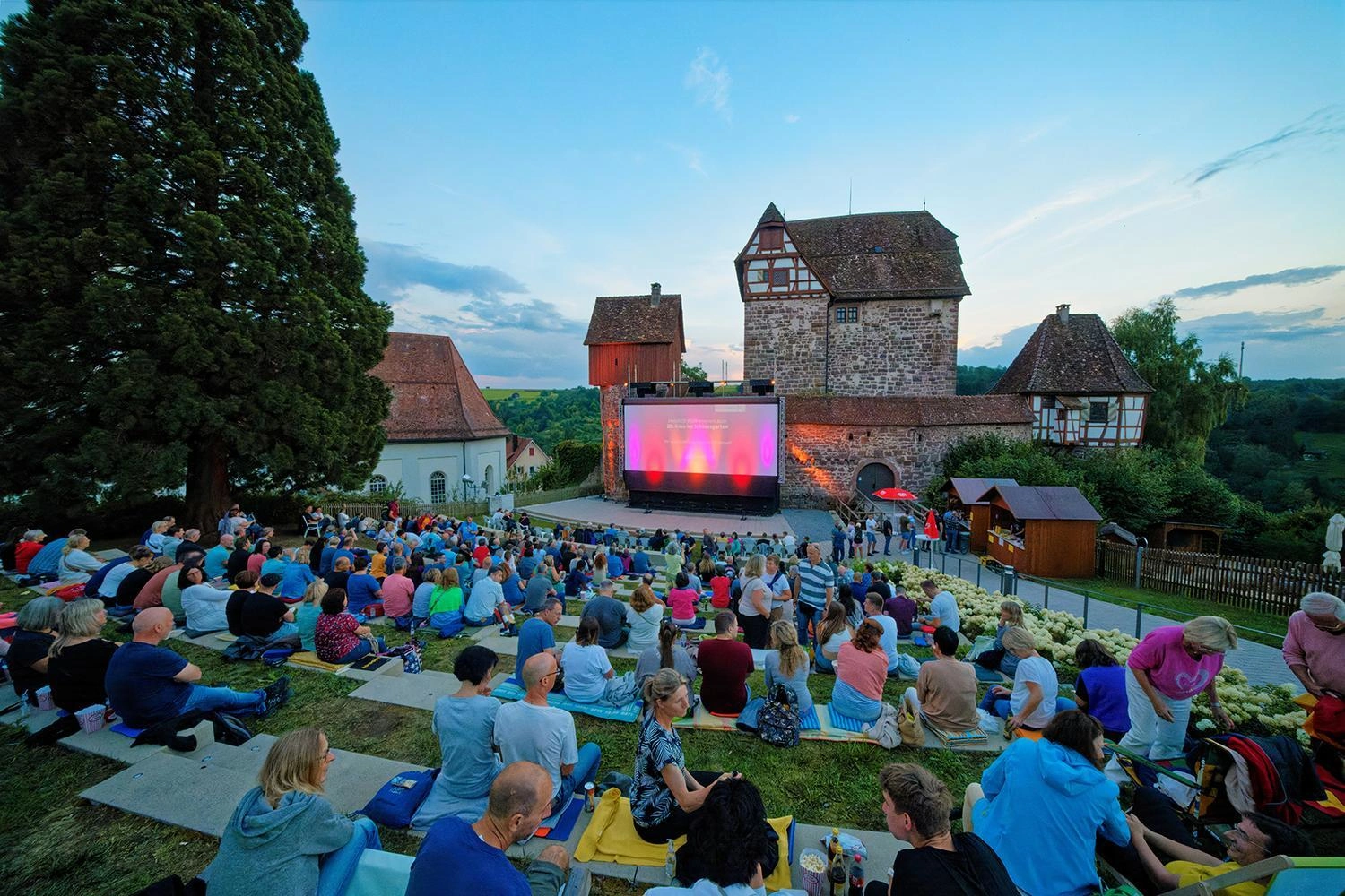 A large cinema screen is set up in front of a historic building. People sit on blankets and chairs in front of it and watch a film at dusk.