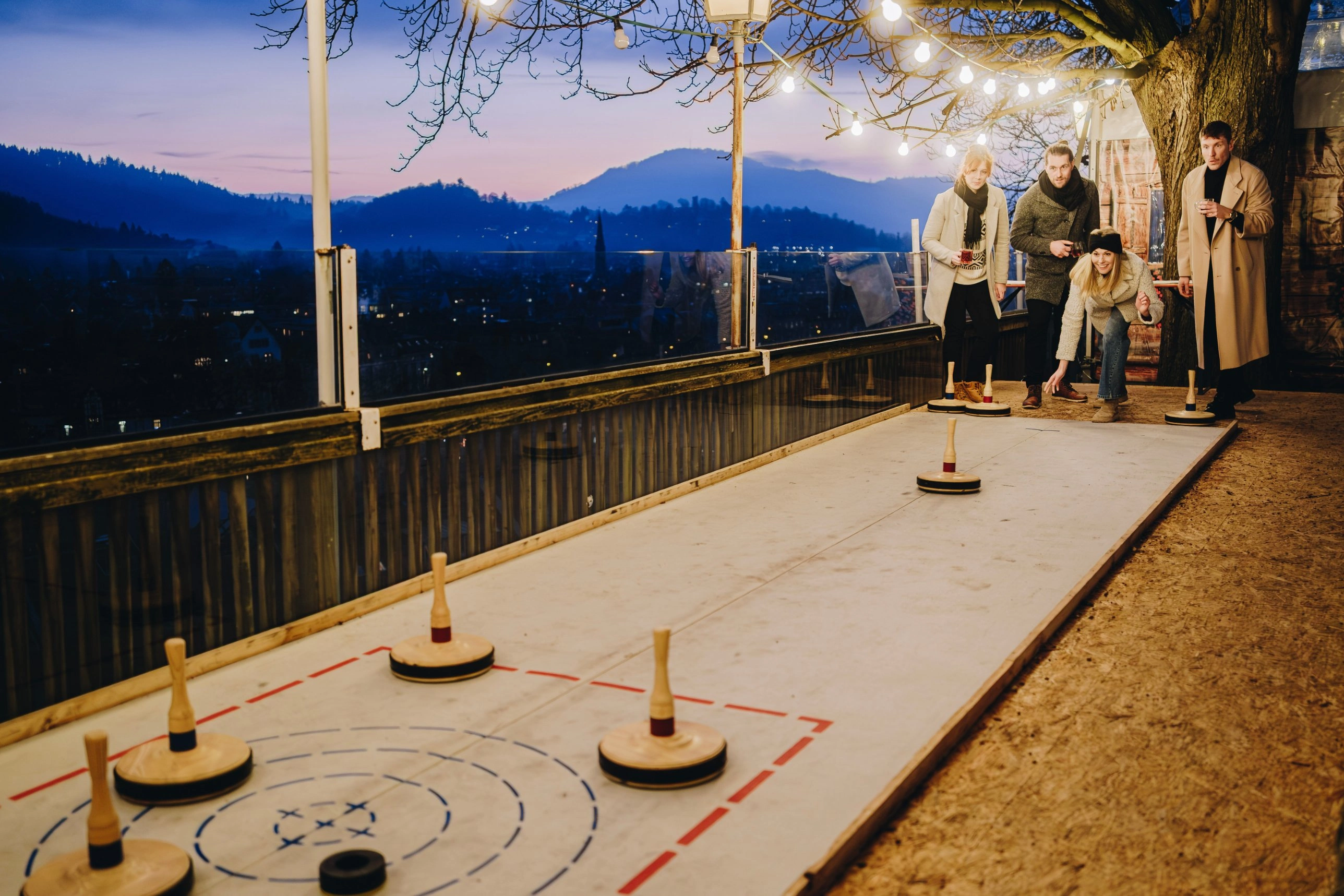 Gäste beim Eisstockschießen auf der Terrasse des Kastaniengartens in Freiburg, dahinter die Altstadt mit Münster in der Abenddämmerung