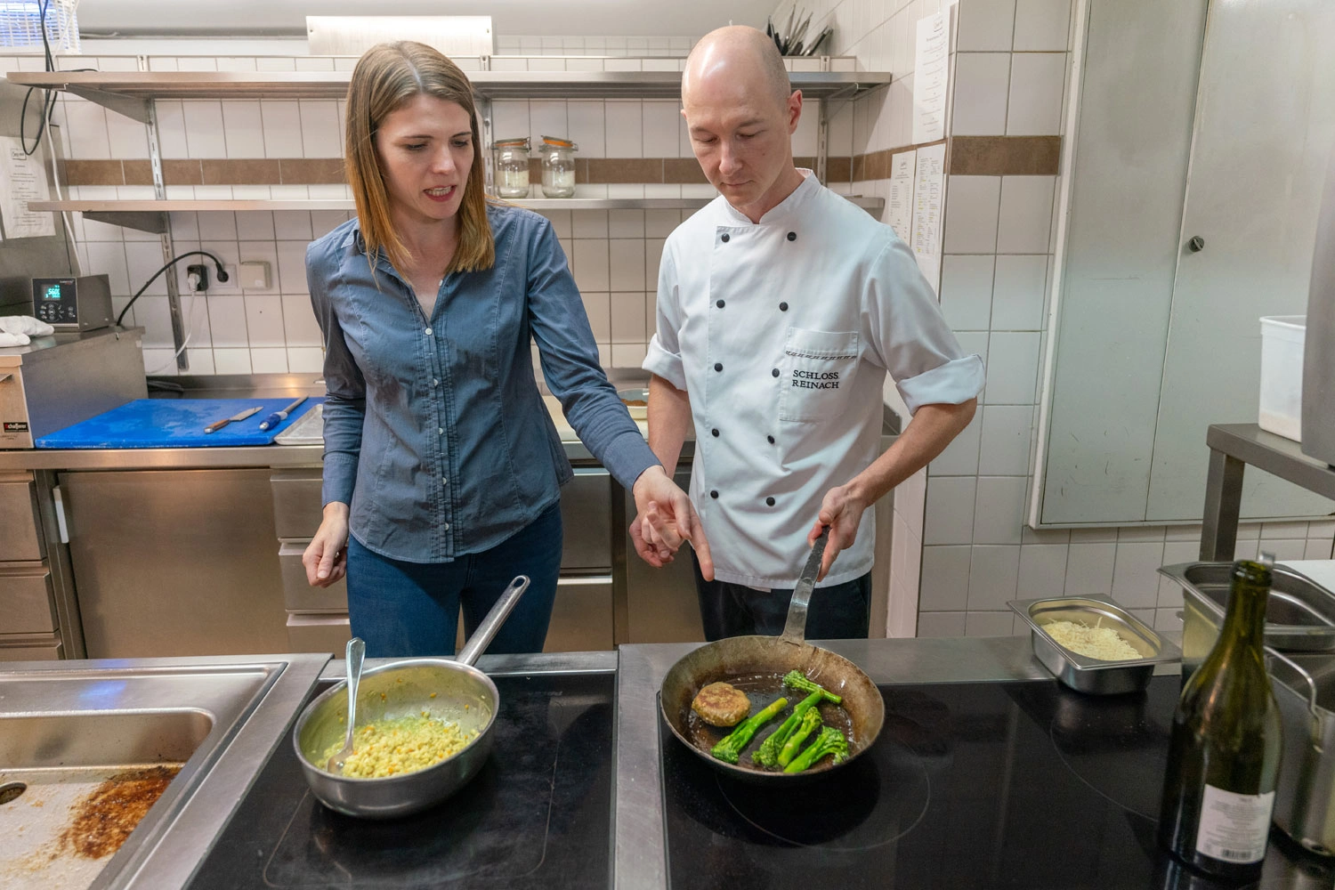 A man and a woman stand together at a stove and discuss the dish in the pan.