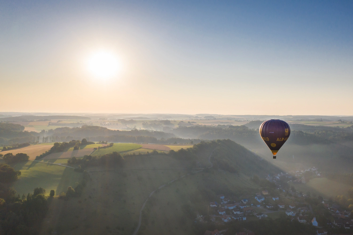 A hot air balloon floats over a vast hilly landscape with fields, forest, and a small village in the valley at sunrise.