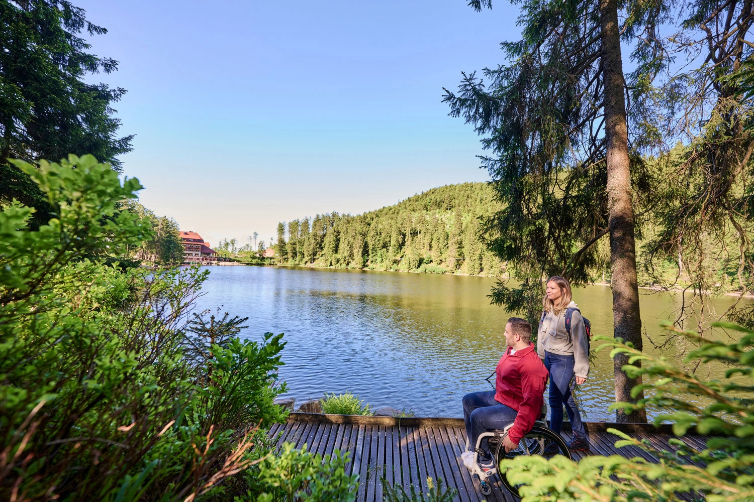 Zwei Menschen genießen den Blick auf einen ruhigen See, umgeben von Wald, auf einem hölzernen Steg im Grünen.