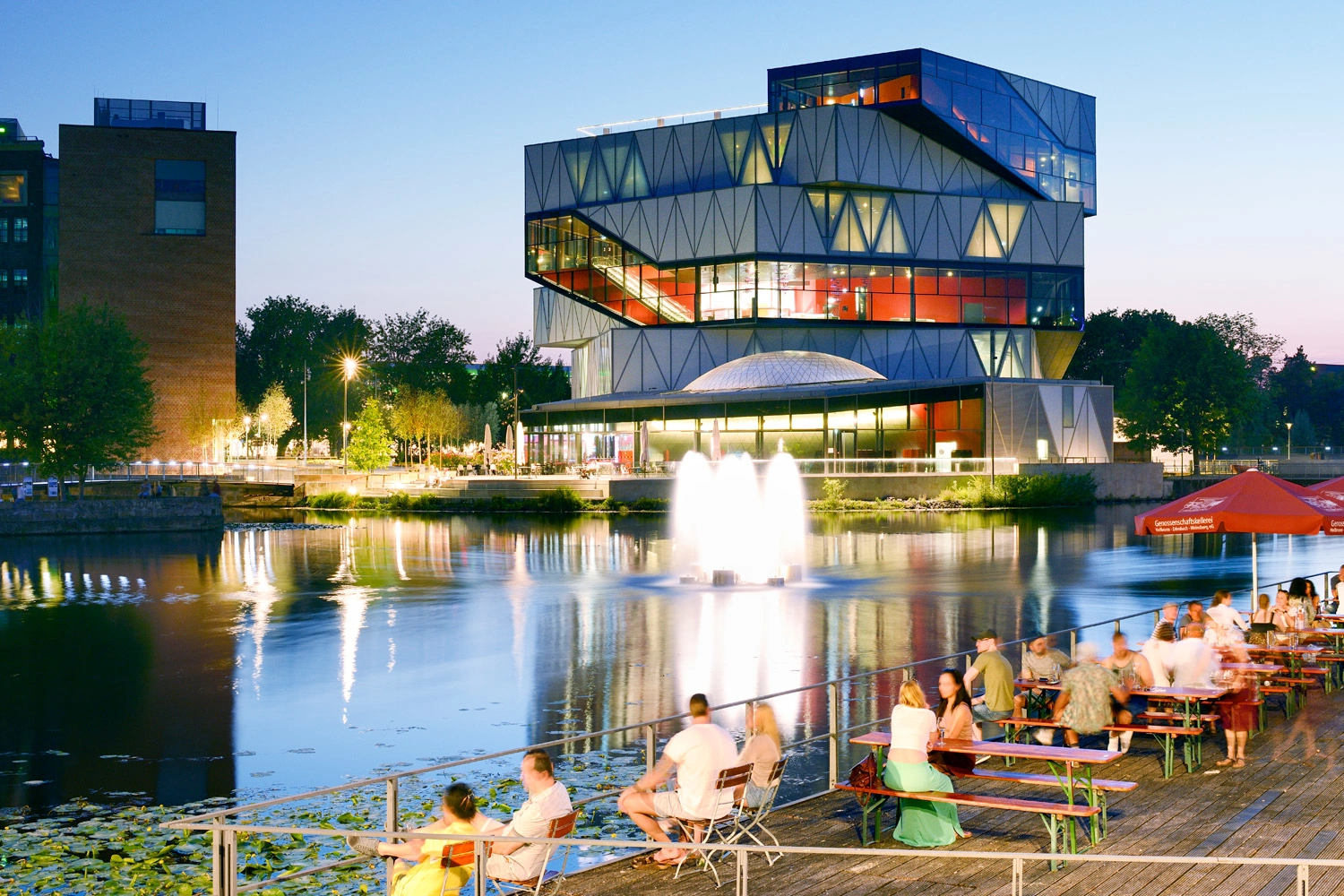 Menschen sitzen auf einer Plattform an einem Gewässer und blicken auf die beleuchtete Fassade der experimenta bei Dämmerung