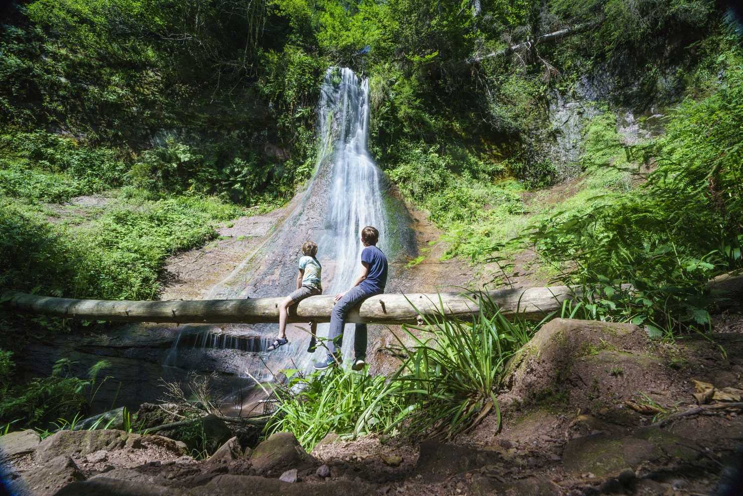 Zwei Wandernde sitzen auf einem Baumstamm vor einem Wasserfall inmitten eines grünen Walds im Schwarzwald.