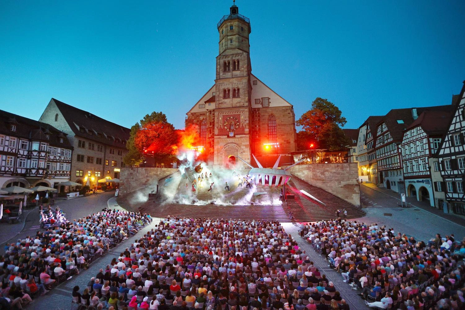 Viele Menschen sitzen auf einem Marktplatz und verfolgen eine Theateraufführung auf einer Treppe. 